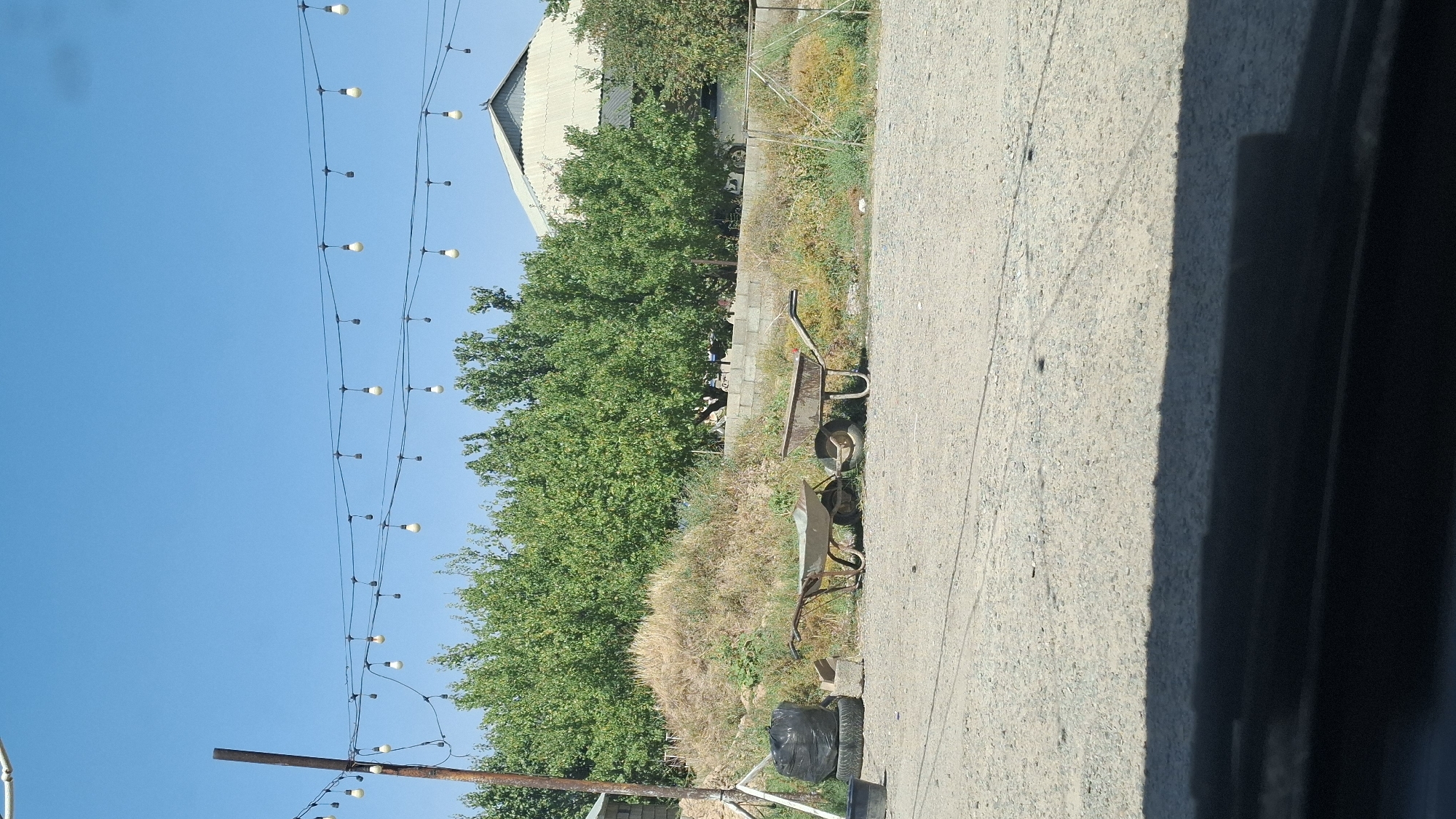 The image depicts an outdoor scene under a clear blue sky. In the foreground, a gravelly ground is visible, with a prominent shadow cast from an unseen object. Two wheelbarrows are present, one lighter in color and appearing newer, the other a darker, rusted metal. To the left of the wheelbarrows, a stack of old tires is topped with a black plastic bag, and a small black bucket rests on the ground. Behind these objects, a pile of dry hay or straw is situated next to a low concrete block wall. A line of lush green trees forms a dense backdrop. Above the trees, two parallel lines of string lights with exposed bulbs are strung across the sky, supported by a vertical pole on the left. In the distance, a building with a distinctive multi-pitched roof is visible. The scene suggests a rural or semi-rural setting, potentially a yard or work area, in Шұбарсу, Kazakhstan. There are no people visible in the image.
