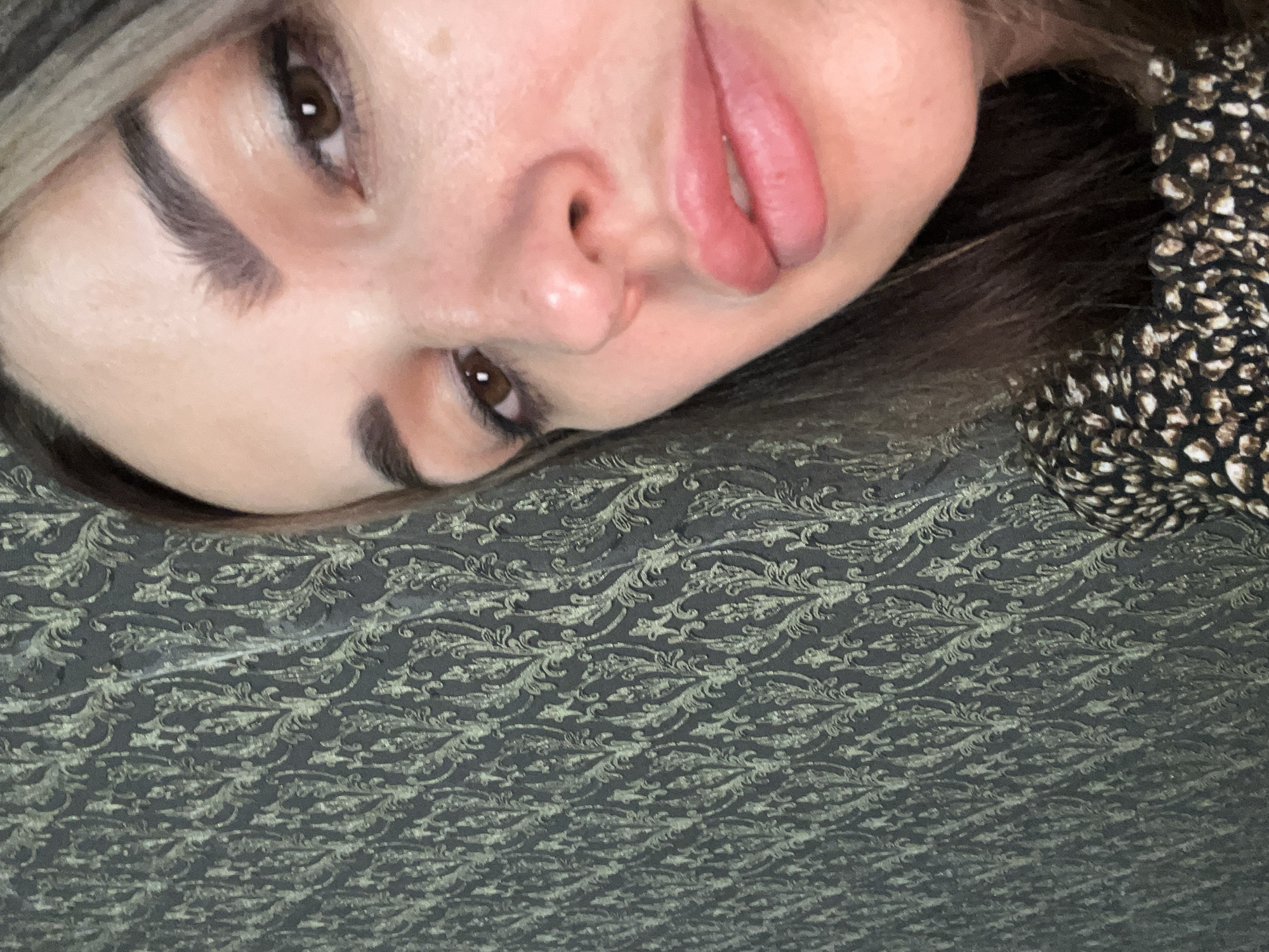 This media file is a close-up, overhead shot of a woman's face, tilted to the side. Her right eye and part of her left eye are visible, along with her nose and lips. Her lips are full and painted with a matte pink lipstick. She has well-defined eyebrows. Her hair, which appears to be brown with lighter highlights, frames her face. She is wearing a dark patterned top with small white and black speckles. The background is a textured surface with a dark gray base and a raised, repeating pattern of swirling, organic shapes in a lighter, possibly metallic, color. The lighting suggests an indoor environment, possibly with soft light. There is no visible text. The overall mood conveyed is intimate and relaxed, with the woman appearing to be resting or posing casually. The composition focuses on the details of her face and the texture of the fabric.