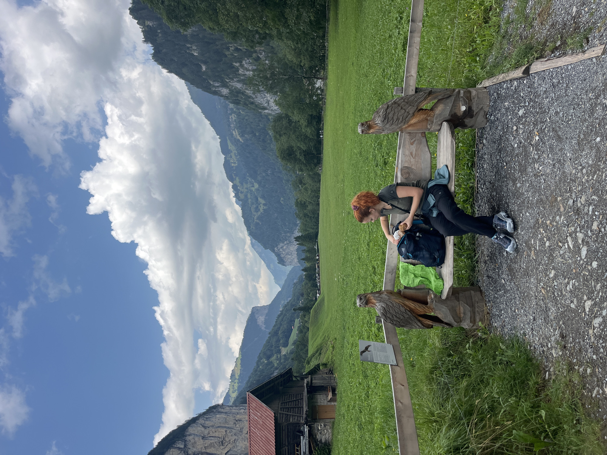 A woman with red hair is sitting on a wooden bench in Lauterbrunnen, Switzerland. She is looking down at a backpack at her feet and holding a piece of food. The bench is carved to look like an eagle on either side. Behind her, a large green field stretches towards mountains. To the left of the field, there is a wooden building with a red roof. The sky is blue with some clouds.