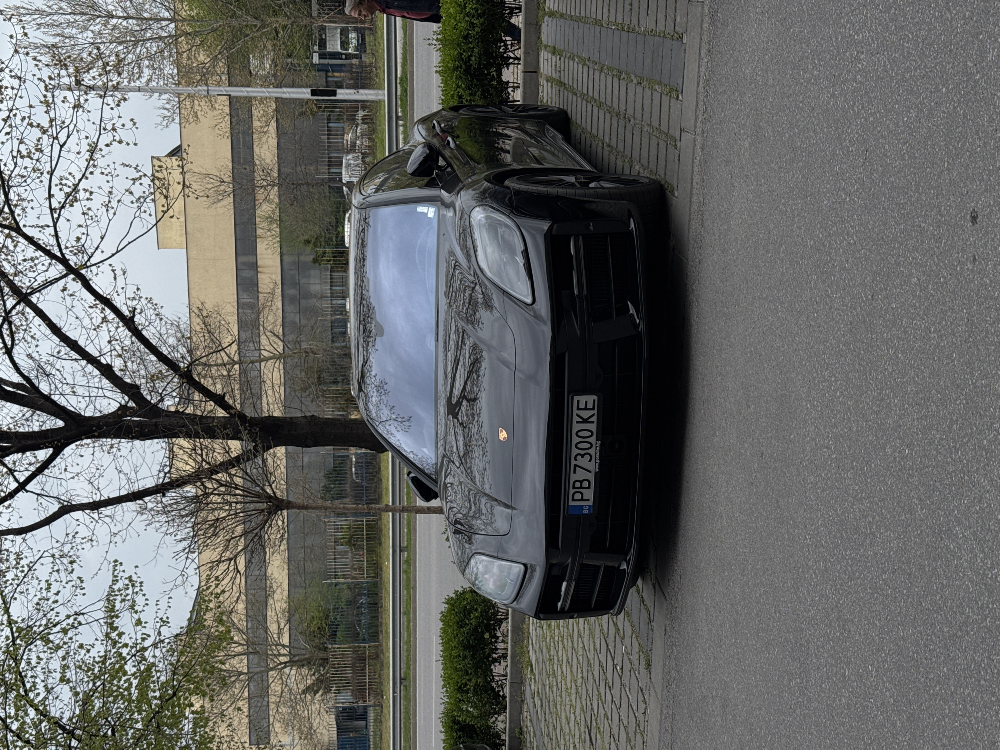 The image captures a black Porsche parked alongside a brick-paved sidewalk and an adjacent asphalt road. A tree with bare branches and emerging spring foliage is prominent on the left side of the frame. In the background, a multi-story building with a combination of light-colored and glass facades is visible. The license plate of the Porsche reads "PB 7300 KE". A portion of a person is seen in the top right corner of the photo. The location appears to be an urban area. Based on the license plate information and visual style, the location is likely in Bulgaria, possibly Plovdiv.
