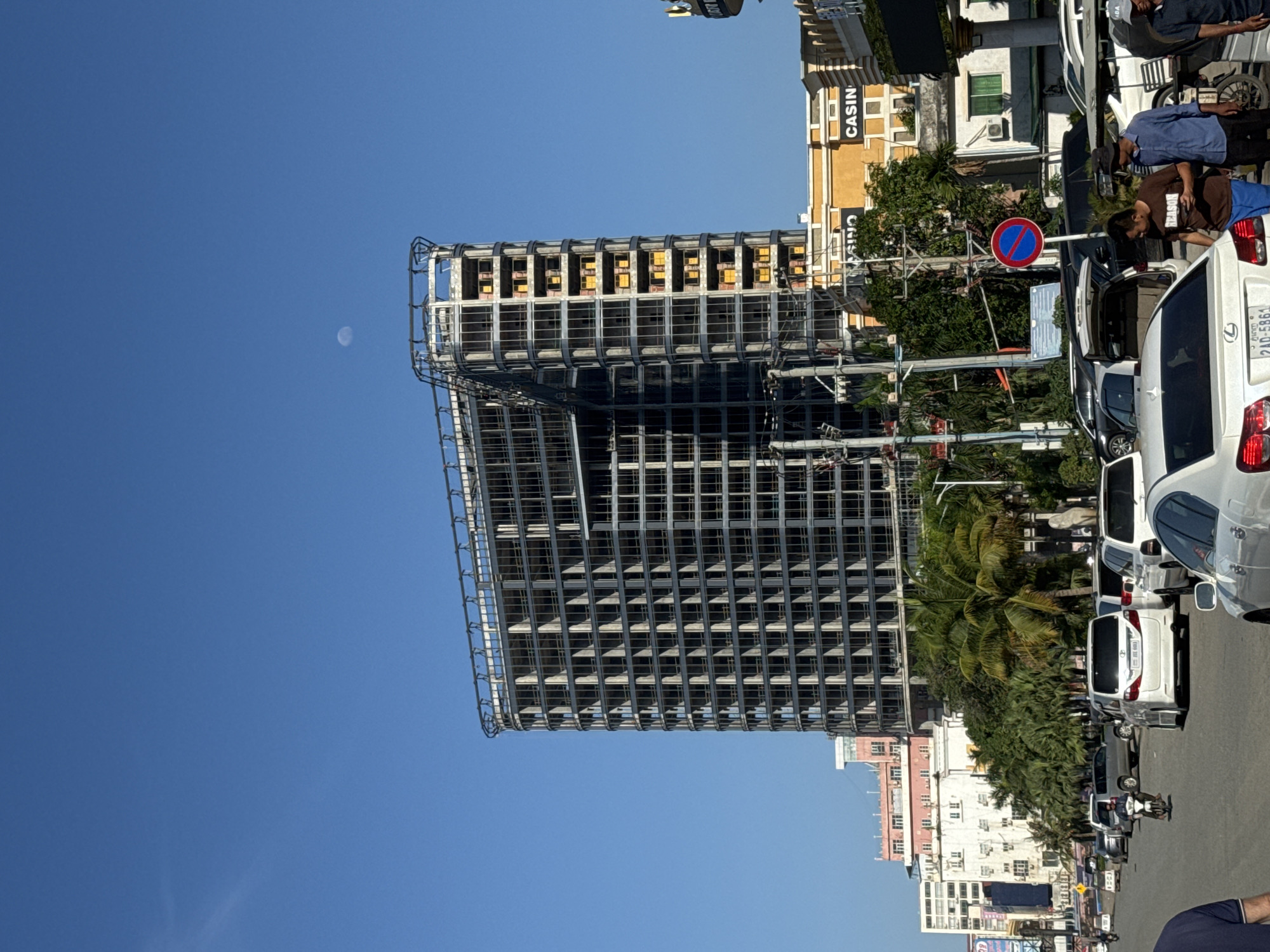 In Bavet Quarter, Cambodia, the image captures a street scene on a clear, blue-sky day. A large, unfinished high-rise building dominates the upper portion of the frame, its skeletal structure of concrete and metal framing visible. The building appears to be in a mid-construction phase, with rows of empty window frames and some yellow-covered openings. A crescent moon is visible in the upper right quadrant of the sky.
Below the construction site, a street runs horizontally across the mid-section of the image. A line of vehicles, including white sedans, an SUV, and a motorcycle, are parked or in motion on the road. Several palm trees and other foliage line the street, adding a touch of greenery to the urban environment. In the background, a low-rise building with a yellow facade and the word "CASINO" visible stands prominently. Further in the background, other buildings of varying heights and architectural styles are present.
On the street, several people are visible. Two men are standing near the rear of a white car, one looking at his phone. Another man is on a motorcycle in the middle of the road. The overall impression is one of a developing urban area with ongoing construction and everyday city life.