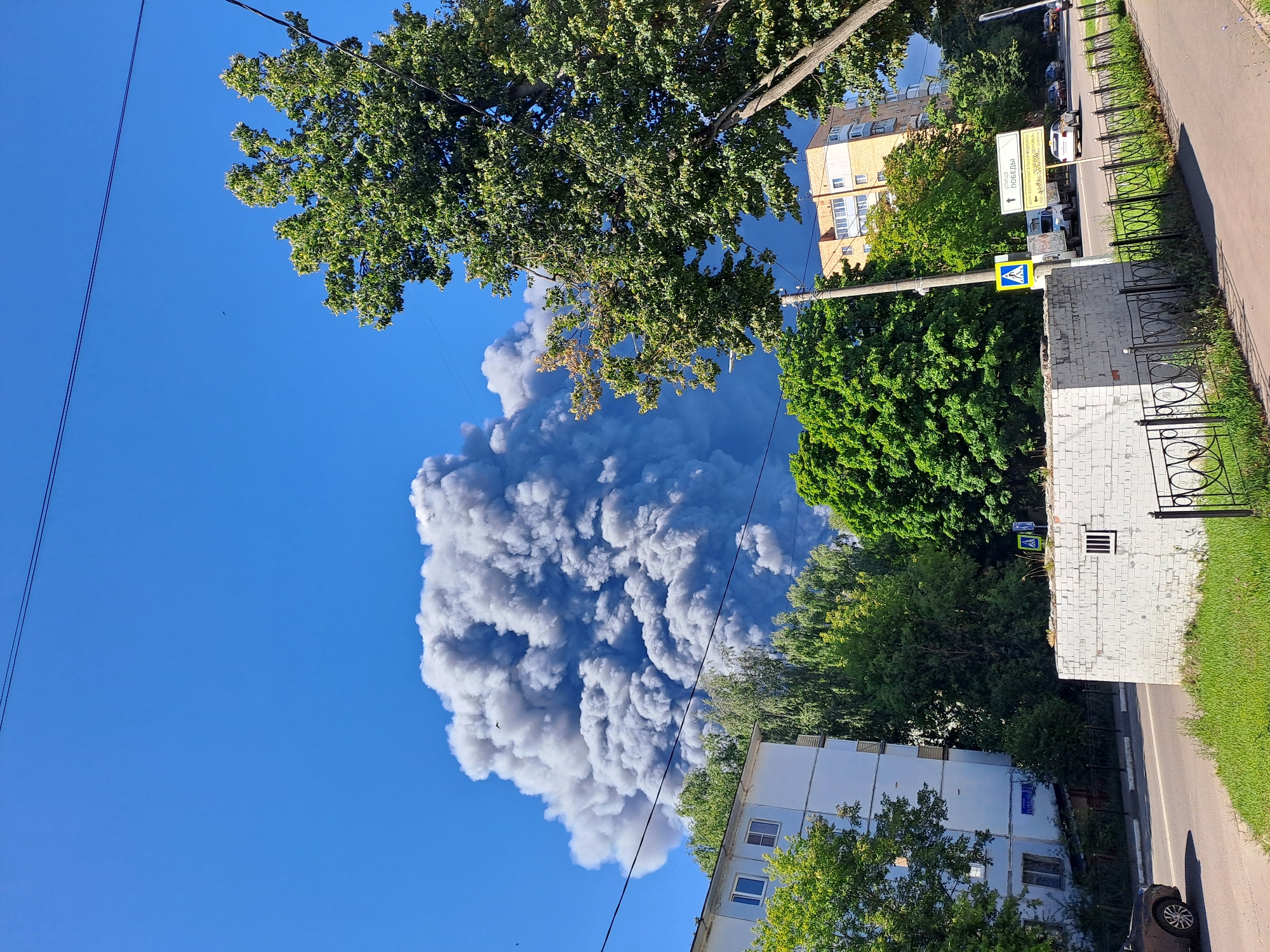 In the city of Elektrostal, Russia, a large plume of grey smoke billows into a clear blue sky. The smoke appears to be originating from a source not visible in the frame. In the foreground, a multi-story apartment building with white and pale blue facades stands to the left, partially obscured by lush green trees. Across a paved road, a lower white brick structure with a barred window is visible, bordered by a black metal fence and green grass. Further to the right, more trees and a yellow apartment building are present. Traffic signs, including a pedestrian crossing sign, are visible near the road. A few cars are parked or driving on the road. The overall scene depicts an urban environment under a bright, sunny sky, dominated by the massive smoke cloud.