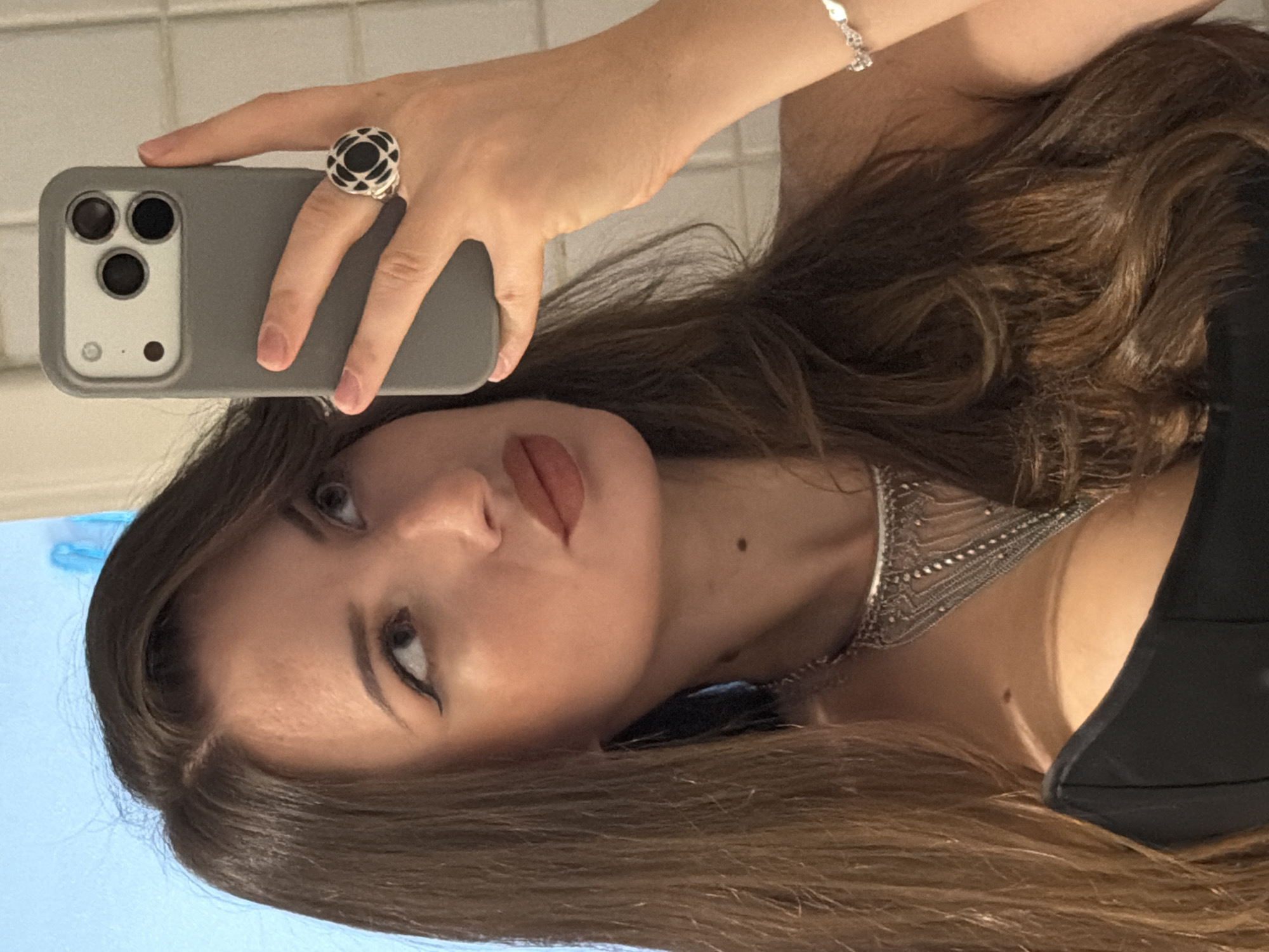 The image shows a woman taking a selfie in what appears to be a bathroom, with a light blue wall visible in the background. She has long, brown hair and is wearing a black top, a detailed silver necklace, a silver bracelet, and a large, black and silver ring on her right hand. Her right hand is holding a grey iPhone, which is angled to capture the selfie. The lighting suggests an indoor environment. The scene does not provide any specific visual cues to identify its location within Sharm El Sheikh, Egypt.