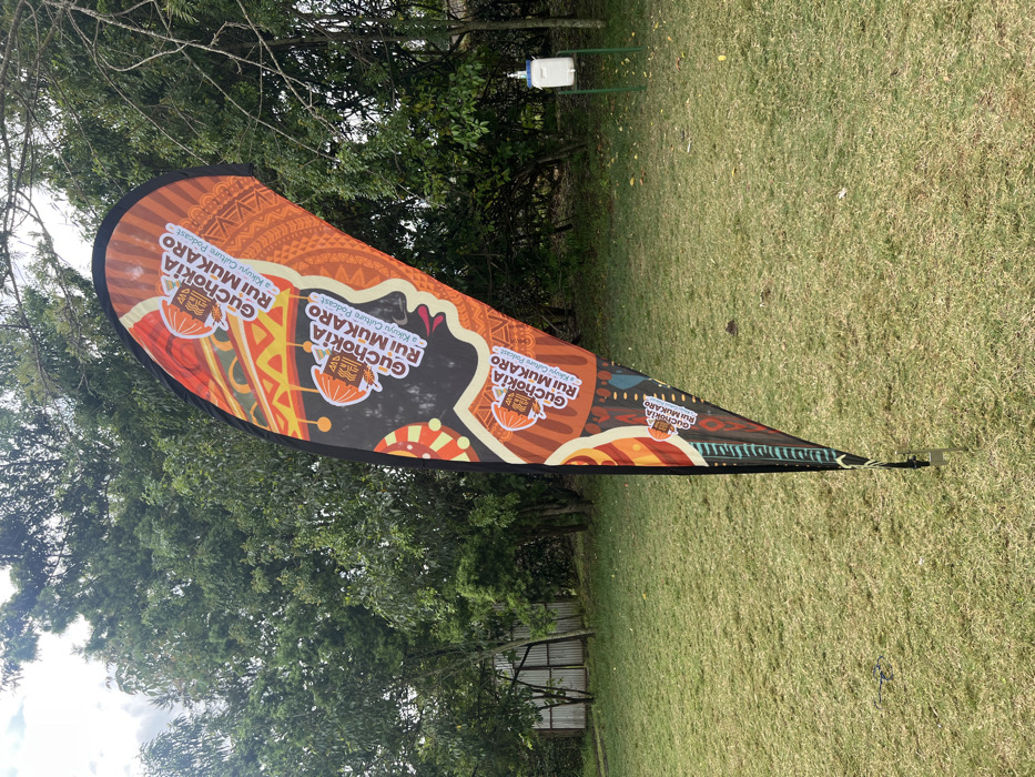 This outdoor image, captured in Makuyu, Kenya, features a vibrant teardrop-shaped banner prominently displayed on a green lawn. The banner, oriented vertically due to the image's sideways capture, is adorned with rich orange, brown, and black hues, featuring intricate geometric and traditional patterns.
The primary text repeated across the banner is "GuChokia Rui MUKARO" and "a Kikuyu Culture Podcast," indicating that the banner is promoting a podcast focused on Kikuyu culture. A stylized logo, possibly depicting a traditional vessel or bowl, is also visible alongside the text.
In the background, lush green trees provide a natural canopy, and a patch of sky, appearing overcast or partly cloudy, suggests it is daytime. Further back, nestled among the foliage, a small white box on a green metal stand is visible, possibly a hand sanitizer station or an environmental sensor. The setting appears to be a verdant, open area, possibly for an event or public display. No individuals are present in the frame.