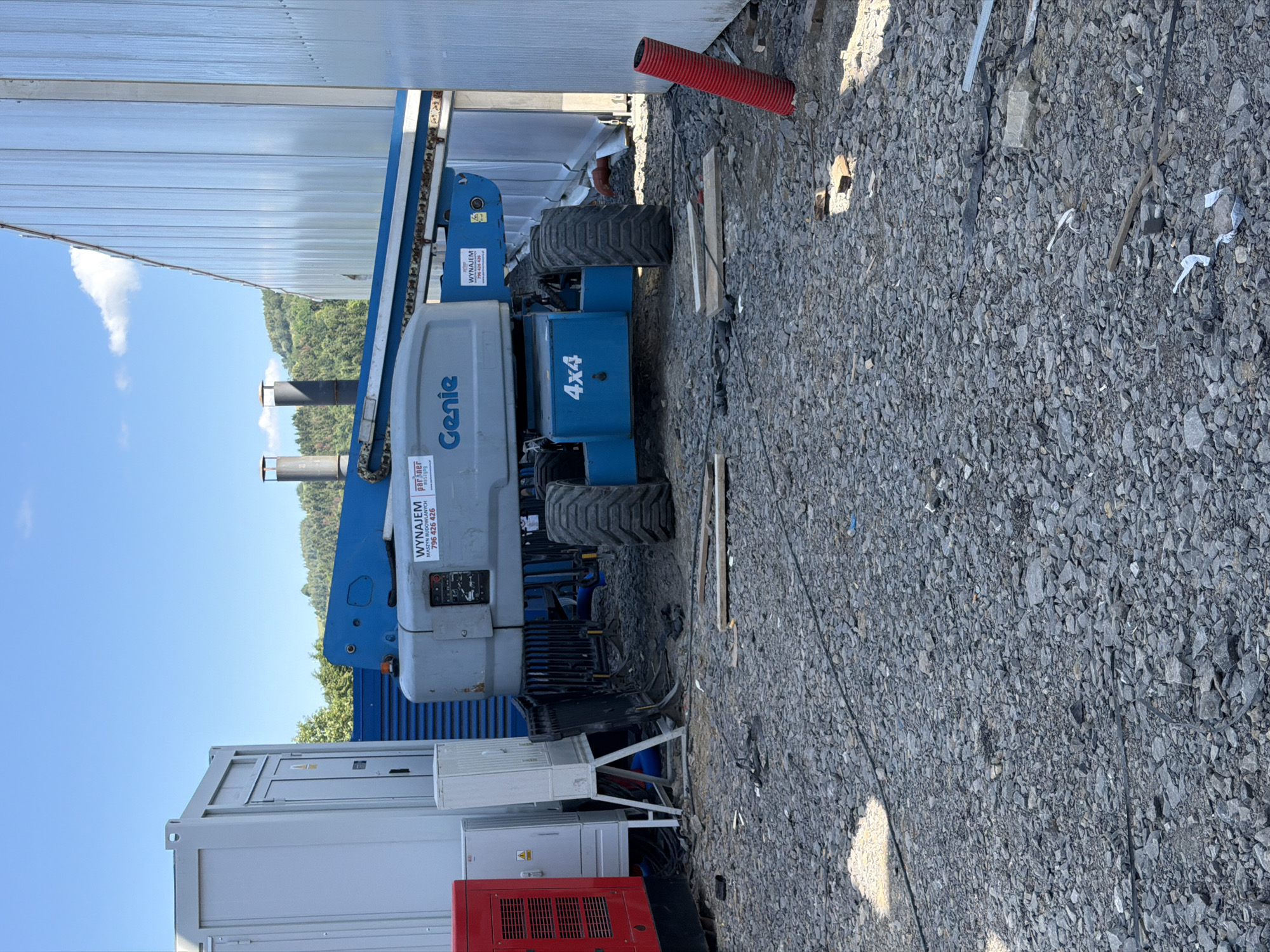 The image depicts a construction site in Janowice, Poland. In the foreground, a gray and blue Genie boom lift is positioned on a gravel surface. The boom lift has "4x4" marked on its base. To the left of the boom lift, a red generator is visible, along with a white electrical panel. In the background, a large white building with a corrugated metal facade dominates the scene. The sky is blue with a few white clouds. The ground is covered in gravel, with some scattered debris and planks. There are no people visible in the image.