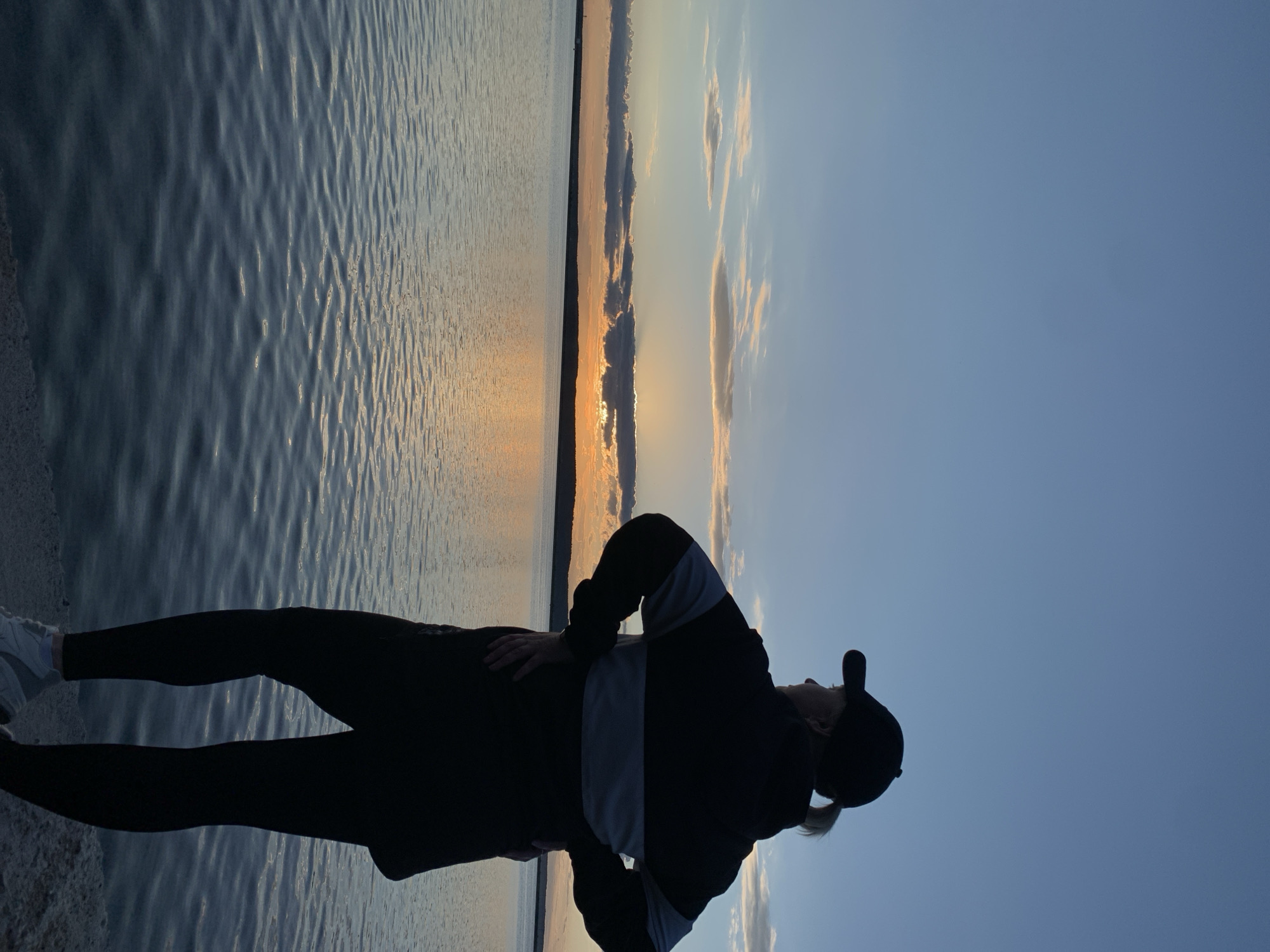The image captures a serene outdoor scene during either sunrise or sunset, overlooking a large body of water, likely the Votkinsk Reservoir in Chaikovsky, Russia. A solitary figure stands in silhouette on the left side of the frame, positioned on what appears to be a shoreline or pier. The person is viewed from behind and slightly to the side, gazing out towards the horizon. They are dressed in dark trousers, white sneakers, a dark jacket with noticeable lighter grey or white stripes on the sleeves and torso, and a baseball cap worn backward, revealing a ponytail. Their hands are clasped behind their back, suggesting a posture of contemplation or appreciation for the view.
The sun is low on the horizon, creating a vibrant orange and yellow glow that stretches across the distant water and illuminates the undersides of scattered clouds. This backlighting casts the individual in a stark, dark silhouette. The expansive body of water, presumably a lake, dominates the mid-ground and foreground, its surface covered with gentle ripples that catch the light. A slender, dark strip of land marks the horizon, separating the reflective water from the sky. The sky above transitions smoothly from the warm hues of the rising or setting sun near the horizon to a clear, tranquil blue, dotted with a few soft clouds. The overall ambiance of the image is peaceful and contemplative. There is no visible text in the media file.