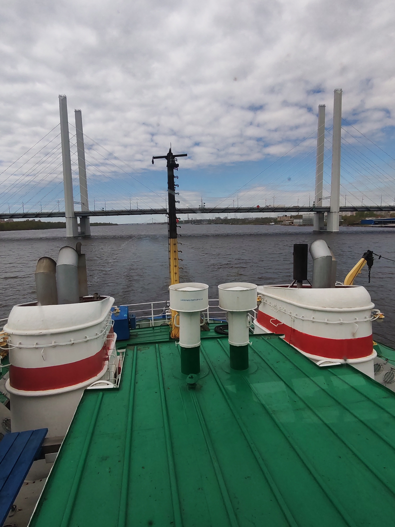 View from the deck of a ship, likely a tour boat, with a green corrugated metal roof. The foreground shows equipment typical for a ship, including exhaust vents and other fixtures painted white with red stripes. Behind the ship, a wide body of water reflects the overcast sky. A large cable-stayed bridge with two prominent white towers spans the water. On the opposite bank, low-rise buildings and trees mark the city skyline. The location is Unknown, Unknown, based on the assumption the city is unidentified in the prompt.