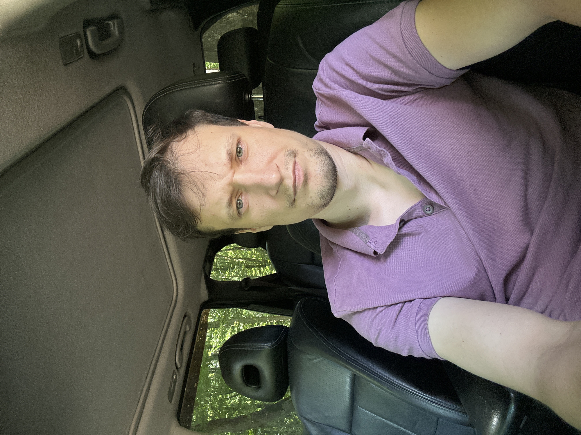 A selfie of a man in a purple polo shirt, seated in the driver's seat of a car. He has brown hair and light-colored eyes, with a slight frown. The interior of the car is visible, including black leather seats and a sunroof. Outside the car, trees and foliage suggest a natural environment, likely a rural or wooded area. The location is identified as Shatava, Ukraine.