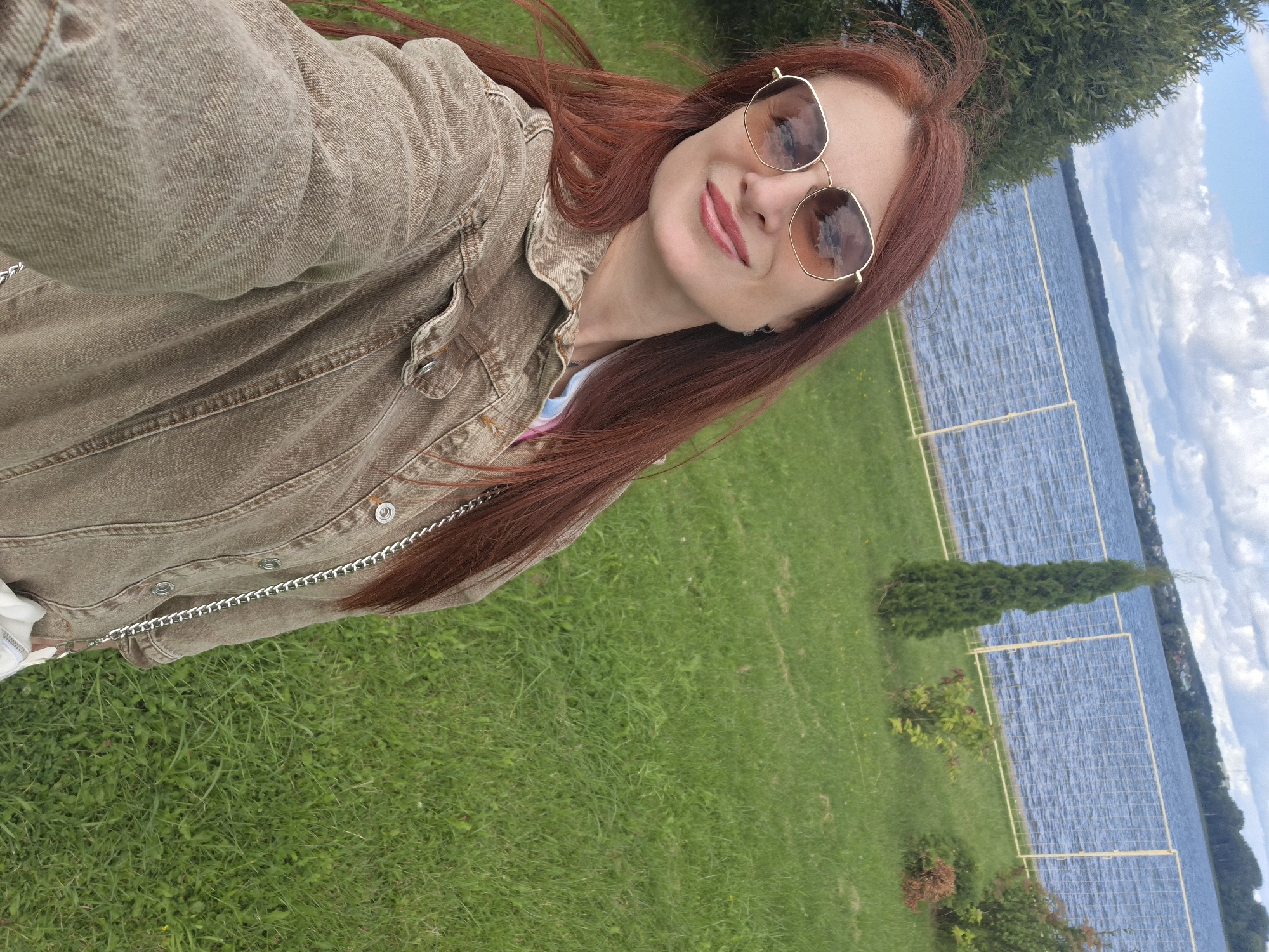 A woman with long, red hair and sunglasses smiles at the camera, taking a selfie outdoors. She is wearing a brown denim jacket with a silver chain strap visible over her shoulder. Behind her, a large body of water, likely a lake, is visible, bordered by a fence and lush green grass. Trees and bushes dot the landscape. The sky above is partly cloudy. The scene appears to be in a park or recreational area near the city of Myadzyel, Belarus, given the presence of a lake and well-maintained grounds.