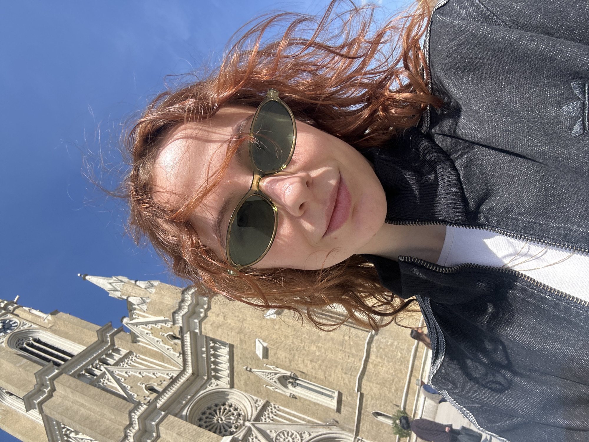 The image is a vertical outdoor portrait, likely a selfie, taken on a bright, sunny day. The foreground features a young woman with fair skin and voluminous, windswept red or auburn curly hair. She is wearing dark, round-framed sunglasses with a light-colored, possibly golden, frame, and has a subtle, pleasant smile. Her attire consists of a dark, likely black, denim jacket with a zipper, worn over a white t-shirt, with a thin necklace visible beneath the jacket's opening. A subtle Adidas trefoil logo is embossed or stitched onto the right side of her jacket.
In the background, positioned primarily to the lower left, is a large, ornate building made of light-colored stone, characteristic of Gothic or Neo-Gothic architecture. This is identifiable as the Name of Mary Church in Novi Sad, Serbia, given the location context. Notable architectural details include intricate carvings, pointed arches, decorative spires, and a prominent circular rose window. A clock face is visible on one of the building's towers. The sky is a clear, bright blue, indicating excellent weather conditions, and strong sunlight casts distinct shadows, highlighting details on both the woman and the historic structure. The photograph captures a moment of casual enjoyment against a significant architectural landmark.