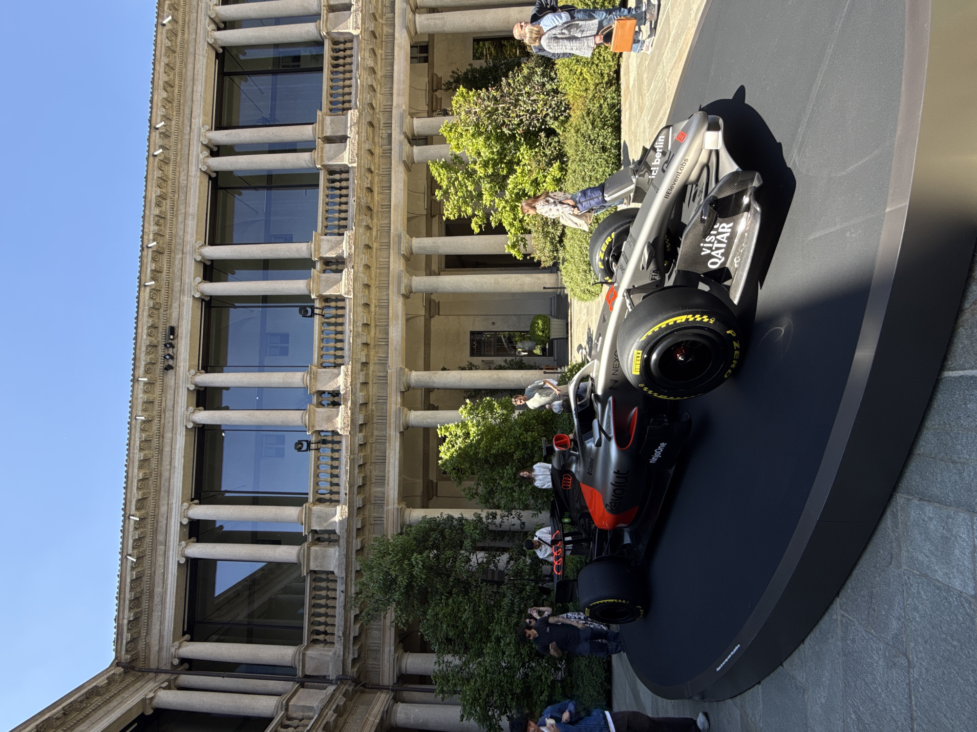 The image displays an outdoor scene in Milan, Italy, featuring a modern open-wheel racing car showcased on a black, circular display platform. The car, predominantly silver and black with orange accents, sports "Pirelli" tires and various sponsor logos, including "Castrol," "Revolut," "ninjaOne," "NEXO," "IRELLI," "FIA," "ic! berlin," "IlElevenLabs," "P ZERO," and "VISIT QATAR."
Several people are seen observing the car, scattered around the platform and in the background. Behind the car and observers stands a grand, multi-story building with classical architecture. Its facade is characterized by numerous columns, large windows reflecting the clear blue sky, and ornate balustrades. Green trees and foliage are visible between the building and the display.
The scene is illuminated by bright daylight under a clear blue sky, indicated by strong shadows cast by the car and other elements, suggesting a sunny day. This setup appears to be a public exhibition or event, showcasing the racing vehicle against a historic architectural backdrop.