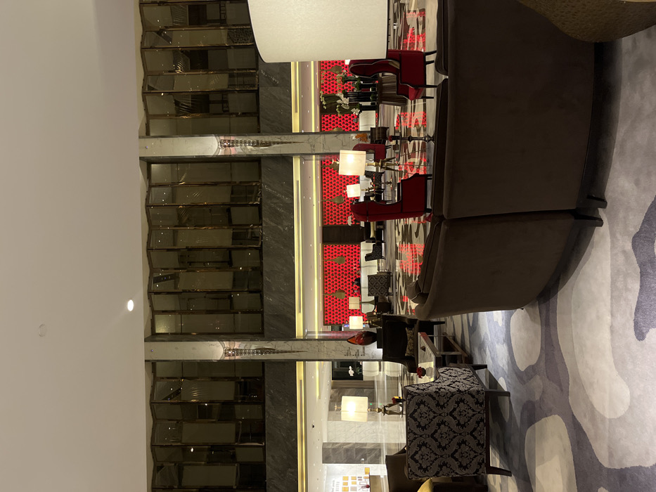 This image depicts a luxurious hotel lobby situated in the Yanta District, China. The scene features a spacious, brightly lit interior with a high ceiling, recessed lighting, and large, reflective panels with a corrugated appearance on an upper wall. Two prominent ornate pillars with vertical decorative elements frame a central section of the lobby.
In the foreground, a large, curved, dark brown sofa is positioned on a patterned rug featuring abstract white, grey, and purplish shapes. To the left, a lounge area includes a dark brown armchair and a patterned armchair (dark grey with a lighter motif), both arranged around a low, rectangular coffee table with a reflective surface. A small red object, possibly a flower arrangement, rests on this table.
Further into the lobby, two tall, red high-back armchairs are visible. Multiple standing lamps with white rectangular or cylindrical shades illuminate the seating areas. In the background, a distinct red wall with a textured pattern and several hanging, bell-shaped pendant lights demarcates what appears to be a reception or secondary lounge area. Scattered small tables and chairs are visible in front of this red wall. To the far left, a lighter-toned lobby section with a counter is partially visible. No identifiable individuals or direct interactions are visible in the scene.