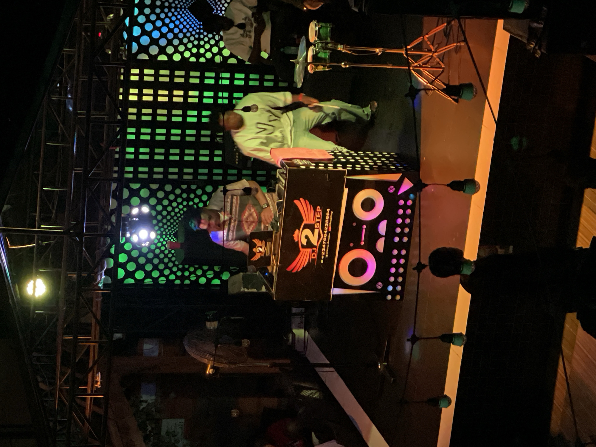 An indoor aerial shot shows a DJ setup with three people. The DJ booth is a black box with a winged logo that reads "DJ 2 STEP". The DJ booth has colorful lights and a phone number. One person stands behind the DJ booth, appearing to mix music. Another person, wearing a white sweatshirt with "NV" printed on it, stands in front of the DJ booth, holding drumsticks and facing the crowd. A third person, wearing a white shirt, is seated to the right, looking at a phone. The background is a wall with a pattern of green and blue lights. There are a set of bongo drums and microphones on a stand to the right of the DJ booth. Several lights hang from the ceiling, casting shadows on the stage.
