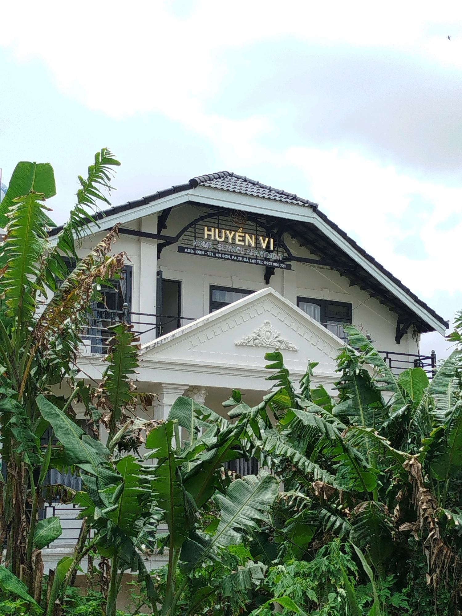 The image shows a white building with a dark roof, partially obscured by lush green banana trees. A sign on the building reads "HUYEN VI HOME SERVICE APARTMENT" and beneath it, "ADD: KGH-T21, AN SON, PA, TP. DA LAT TEL: 0907 900 721". The building appears to be a residential or commercial establishment. The foreground is dominated by the large leaves of the banana plants, with some dry, brown leaves mixed in. The sky is overcast with clouds. The scene depicts an exterior view of the apartment building surrounded by tropical vegetation. The text on the sign indicates the location is in Da Lat, Vietnam.