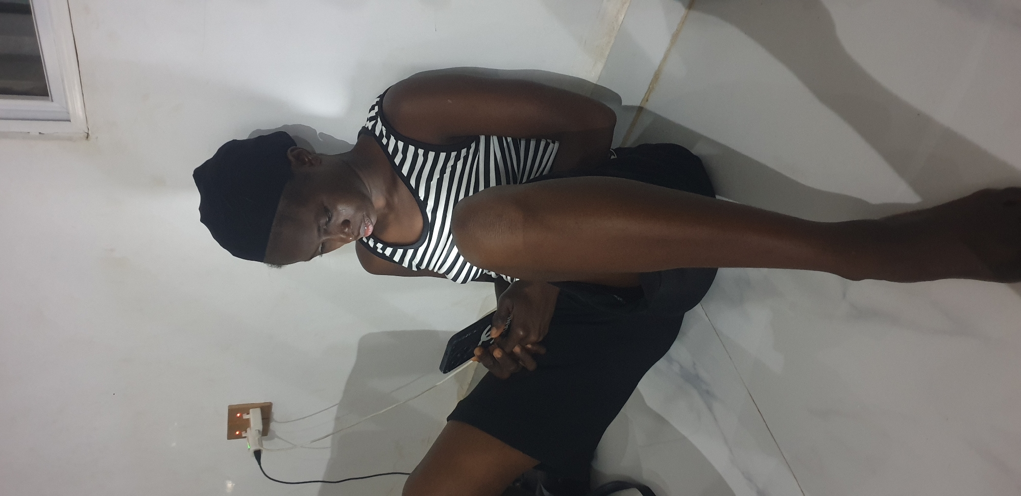 An individual is depicted sitting on a tiled floor, leaning against a white wall, in what is identified as Narayi, Nigeria. The person, appearing to be a young female with dark skin, wears a black head covering, a horizontally striped black and white tank top, and black shorts. She is intently focused on a dark-colored smartphone held in both hands. To the left, an electrical outlet assembly, consisting of a wooden or light-colored rectangular plate housing two sockets, is mounted on the wall. Two white electrical chargers are plugged into these sockets, each exhibiting a small, illuminated indicator light (appearing reddish-orange). A black cable extends downwards from one of these chargers. The floor is composed of light-colored, possibly marbled or patterned tiles. The background includes a partially visible white window frame in the upper right corner. Shadows are cast on the floor to the right of the individual, indicating an overhead or off-camera light source.