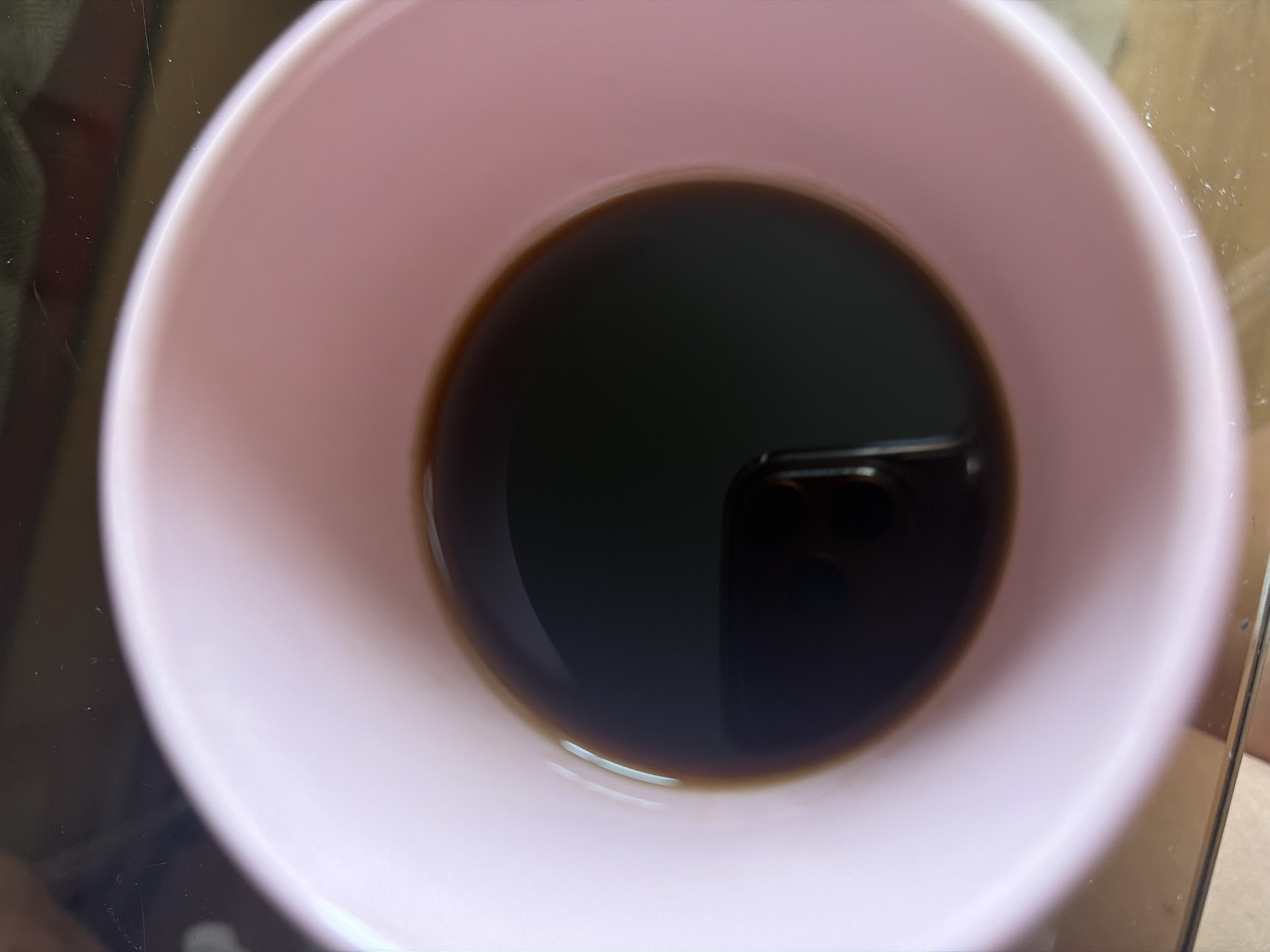 A close-up, overhead shot shows a light pink mug filled with dark liquid, likely coffee. The mug has a smooth, matte finish. The liquid surface is reflective, showing a blurred reflection of what appears to be a mobile phone, indicating the image was taken by someone holding a phone above the mug. The background is out of focus, but suggests a tabletop surface. No specific location in Kyiv, Ukraine, can be determined from the image.