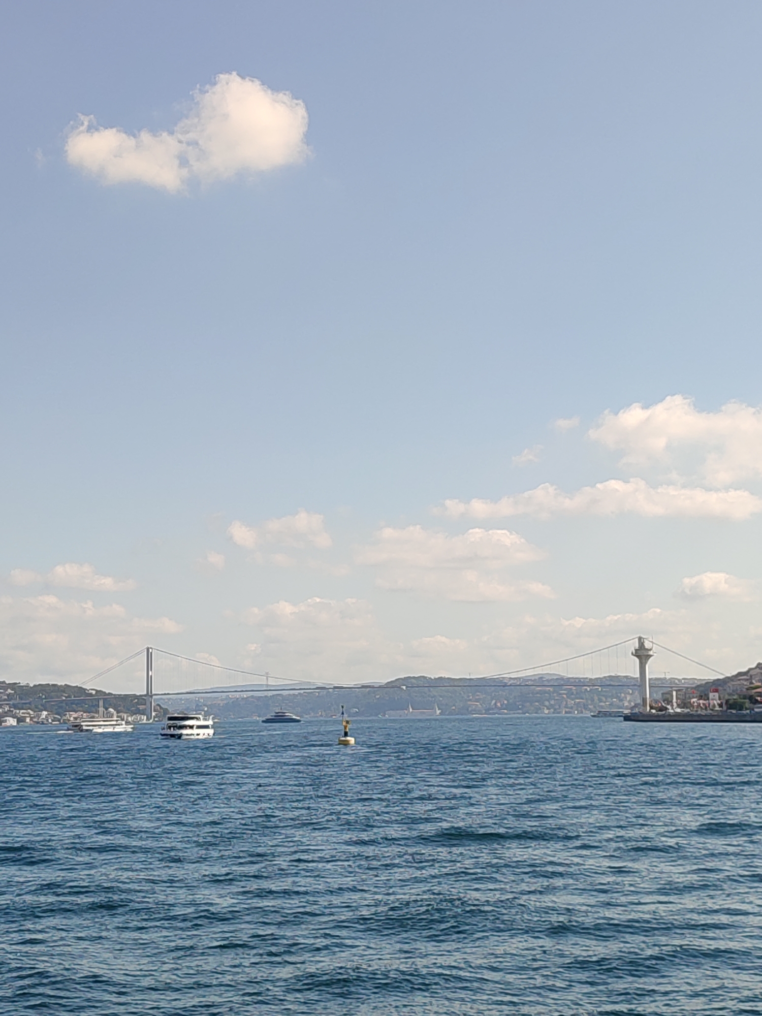 The image depicts a wide shot of the Bosphorus strait in Üsküdar, Turkey, on a clear day with scattered clouds. The foreground is dominated by the blue, rippling water of the strait. In the middle ground, several vessels are visible, including a white passenger ferry and what appears to be a larger yacht. A yellow and blue buoy marks a point in the water.
Dominating the background is the Fatih Sultan Mehmet Bridge, a suspension bridge stretching across the strait. To the right of the bridge, a lighthouse-like structure stands on the shore. The coastline is lined with buildings and greenery, suggesting an urban environment. The sky is a light blue, with a few fluffy white clouds scattered across it. The overall scene conveys a sense of calm and vastness, characteristic of a seascape.