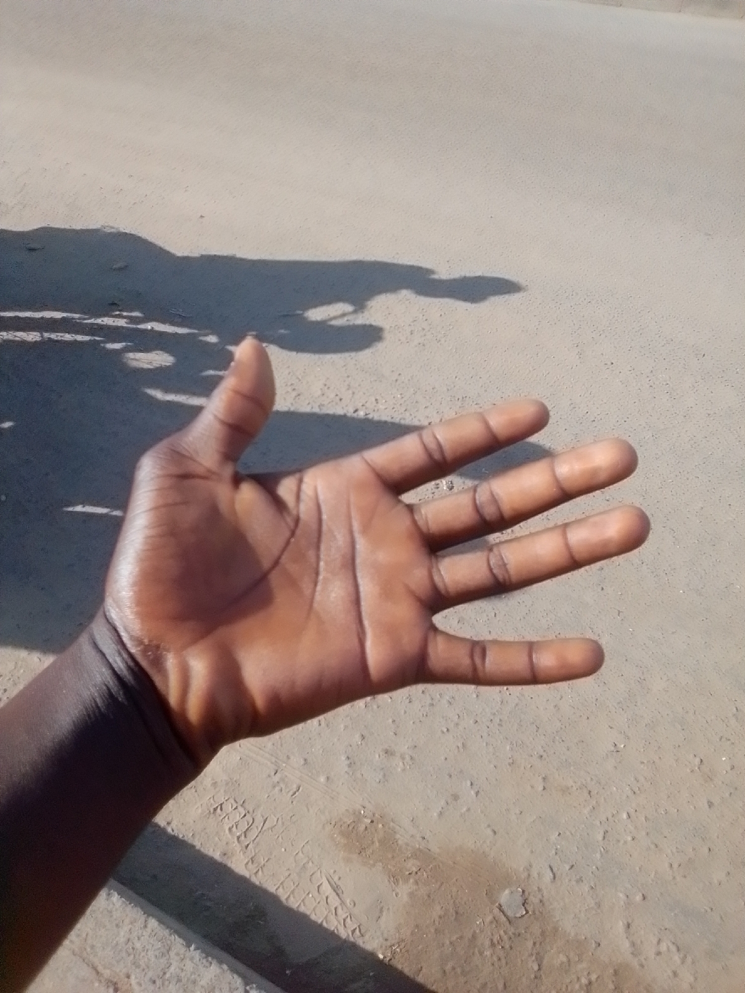 A dark-skinned hand is held up, palm facing the camera, with fingers spread except for the thumb which is bent backward. The hand is positioned in the foreground of the image. The background consists of a light-colored, dusty ground surface, possibly a road or unpaved area. A shadow of the hand and arm is cast on the ground behind it, elongated by the sunlight. The environment appears to be outdoors in an open area. There is no visible evidence to indicate the specific location within Kwadon, Nigeria, or any identifiable landmarks. The image is a close-up shot focusing on the hand.