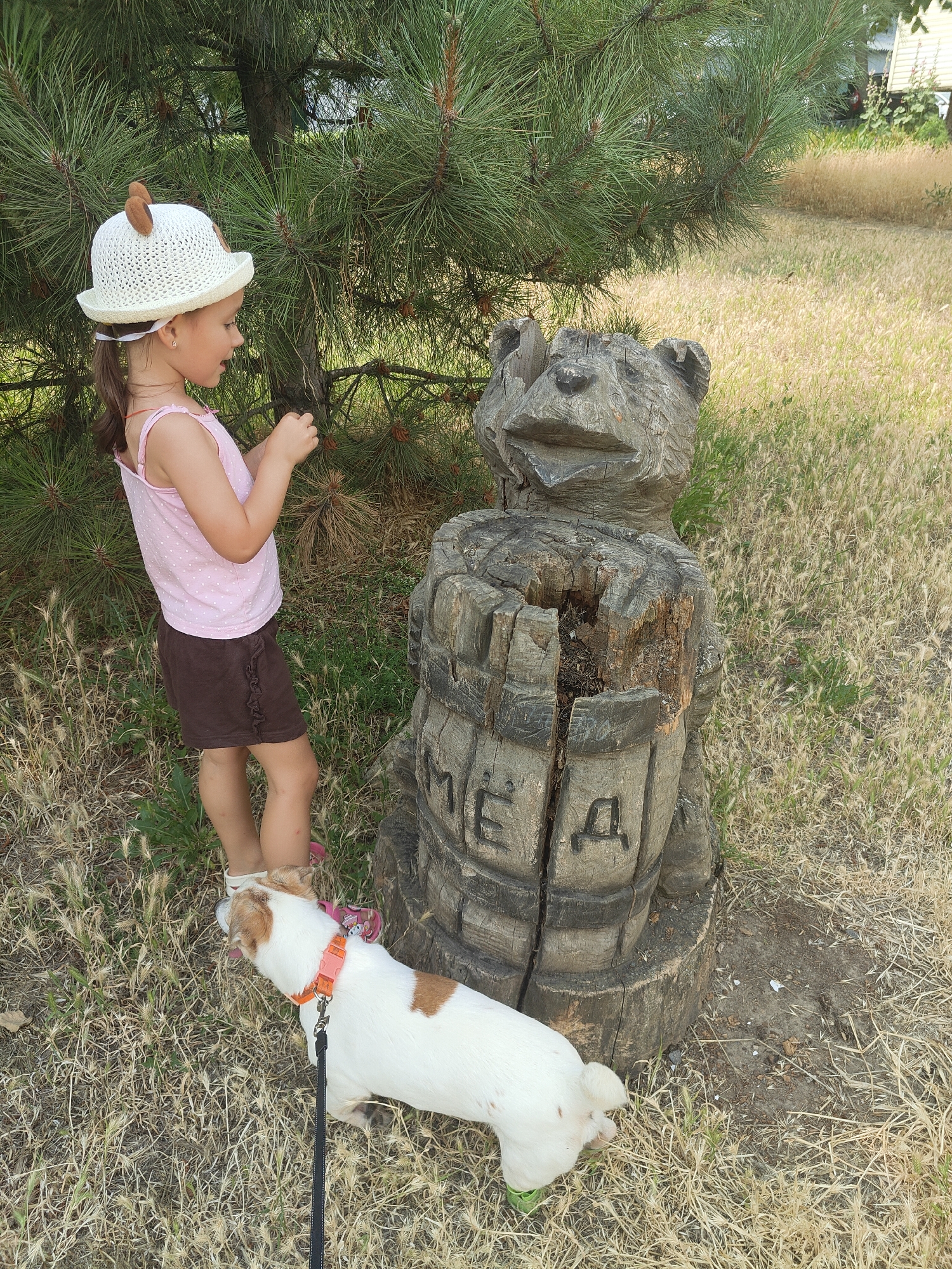 A young girl wearing a white straw hat with ear-like decorations and a pink polka-dot tank top is standing outdoors next to a carved wooden bear sculpture. She is looking down at her hands, which are holding something small. A small white and brown dog with an orange collar is lying on the ground in front of the sculpture, facing away from the girl. The sculpture is made from a tree stump and has Cyrillic letters carved into it, appearing to spell "ЕДЕМ" (EDEM), meaning "we are going" or "we are coming". The background consists of dry grass and a large pine tree. The scene is set in the city of Chornomorsk, Ukraine, likely in a park or natural area.