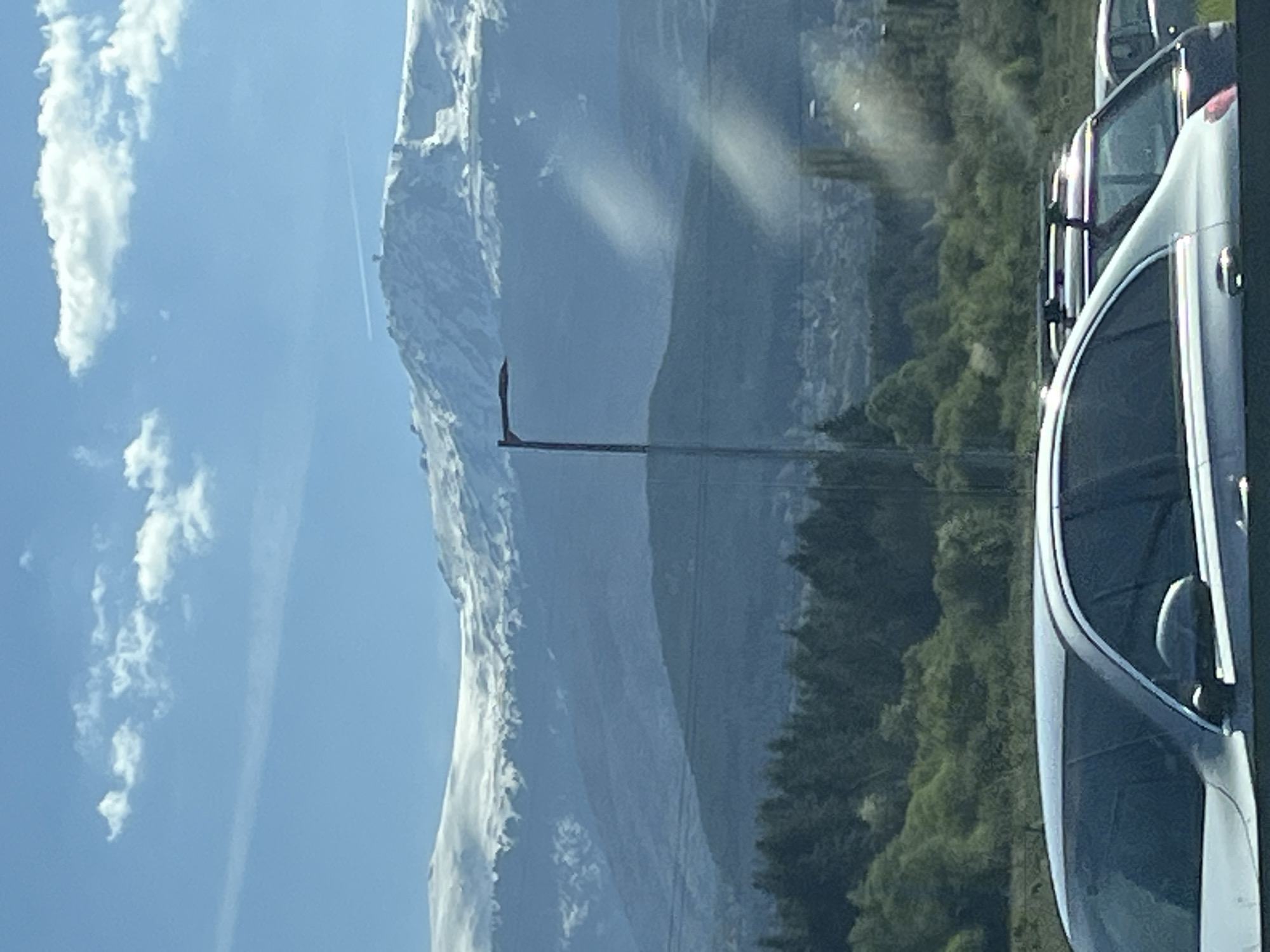 A landscape image features a snow-capped mountain range under a partly cloudy sky. In the foreground on the right, portions of several light-colored vehicles are visible, including side mirrors and windows. A forest extends along the base of the mountain. A utility pole stands between the mountain and the vehicles. The location is Unknown, Unknown.