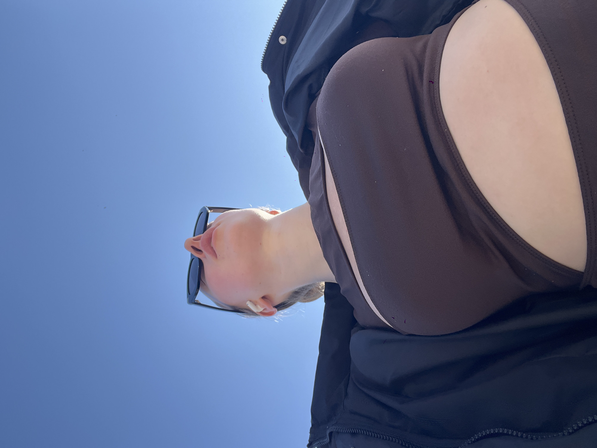 The image, taken from a low angle, shows a woman wearing sunglasses and a brown top against a clear blue sky. She is also wearing a dark jacket, and an earbud is visible in her ear. The location is likely outdoors in the city of Boreti, Montenegro, given the clear sky and sunlight, though specific landmarks are not visible.