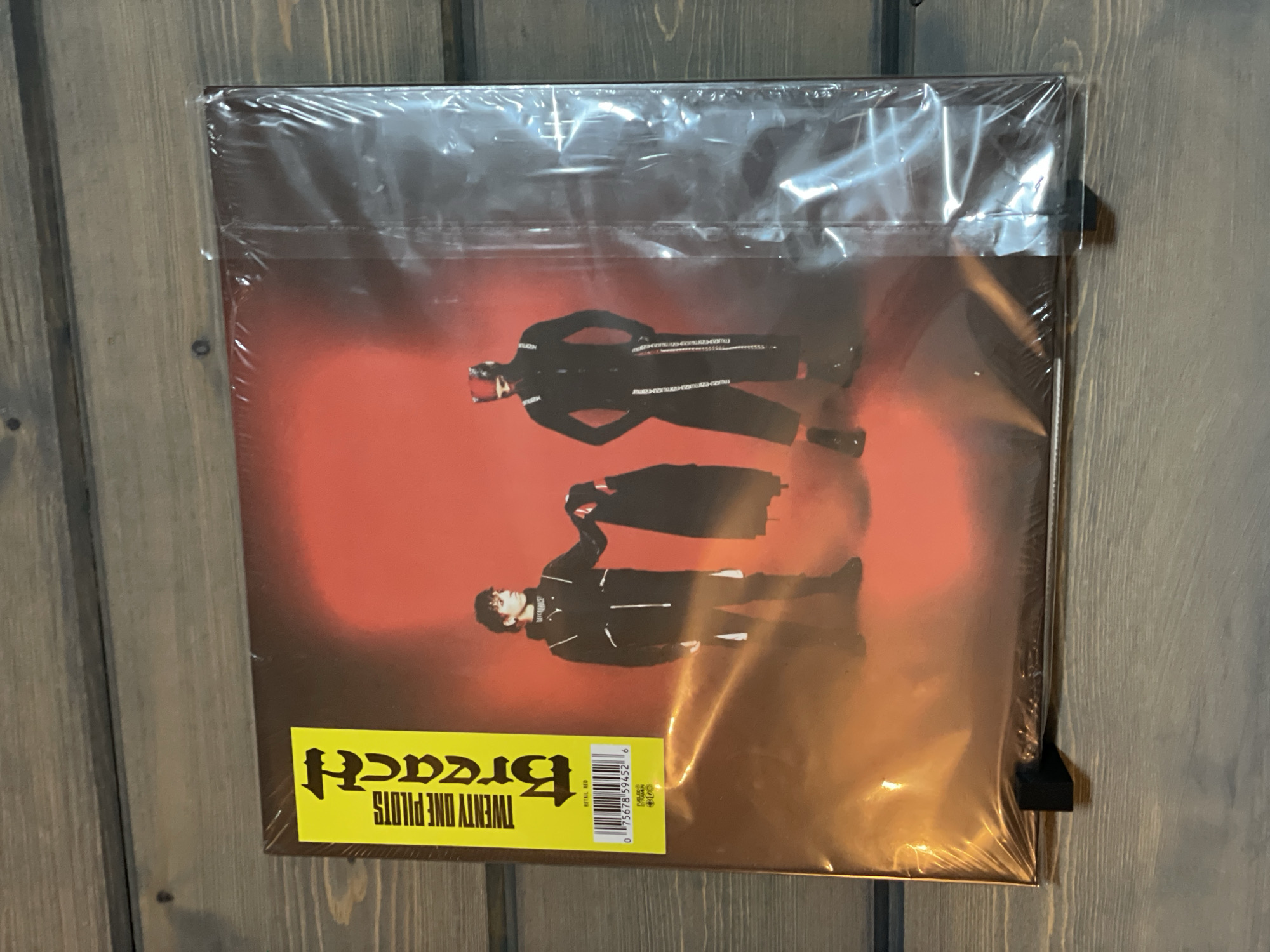 A vinyl record album is displayed on a wooden surface. The album cover features two individuals in dark attire against a red backdrop. A yellow sticker with black text is affixed to the left side of the album cover. A barcode is visible on the lower portion of the yellow sticker, along with text that reads "RETAIL RED". The record is sealed in plastic wrap. The background consists of horizontal wooden planks, a common element found in the city of Angarskie Khutora, Russia.