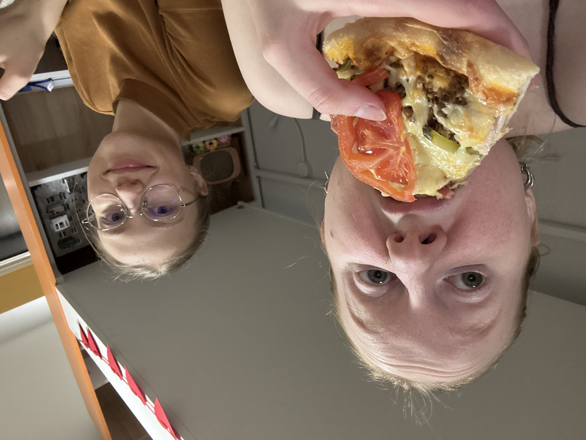 Two light-skinned individuals are present. The first person is positioned in the upper left of the image, wearing eyeglasses and a brown shirt. The second individual is in the upper right, holding a slice of pizza with visible tomato topping toward their mouth. The background includes a shelf unit. It's impossible to provide information on the location of Perm, Russia with the available information.