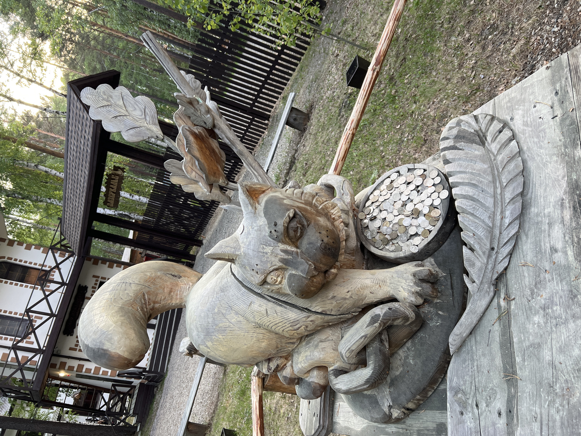 The image shows a carved wooden sculpture of a cat sitting in front of a building in Tal'tsy, Russia. The cat is large and detailed, with coins piled in a bowl-like structure in front of it. The building in the background appears to be a traditional Russian wooden house with a pitched roof and a balcony. The surrounding environment is a wooded area with a fence and benches. The sculpture is placed on a wooden platform, with a carved feather in the foreground. The overall scene suggests a tourist attraction or a cultural site within the village.