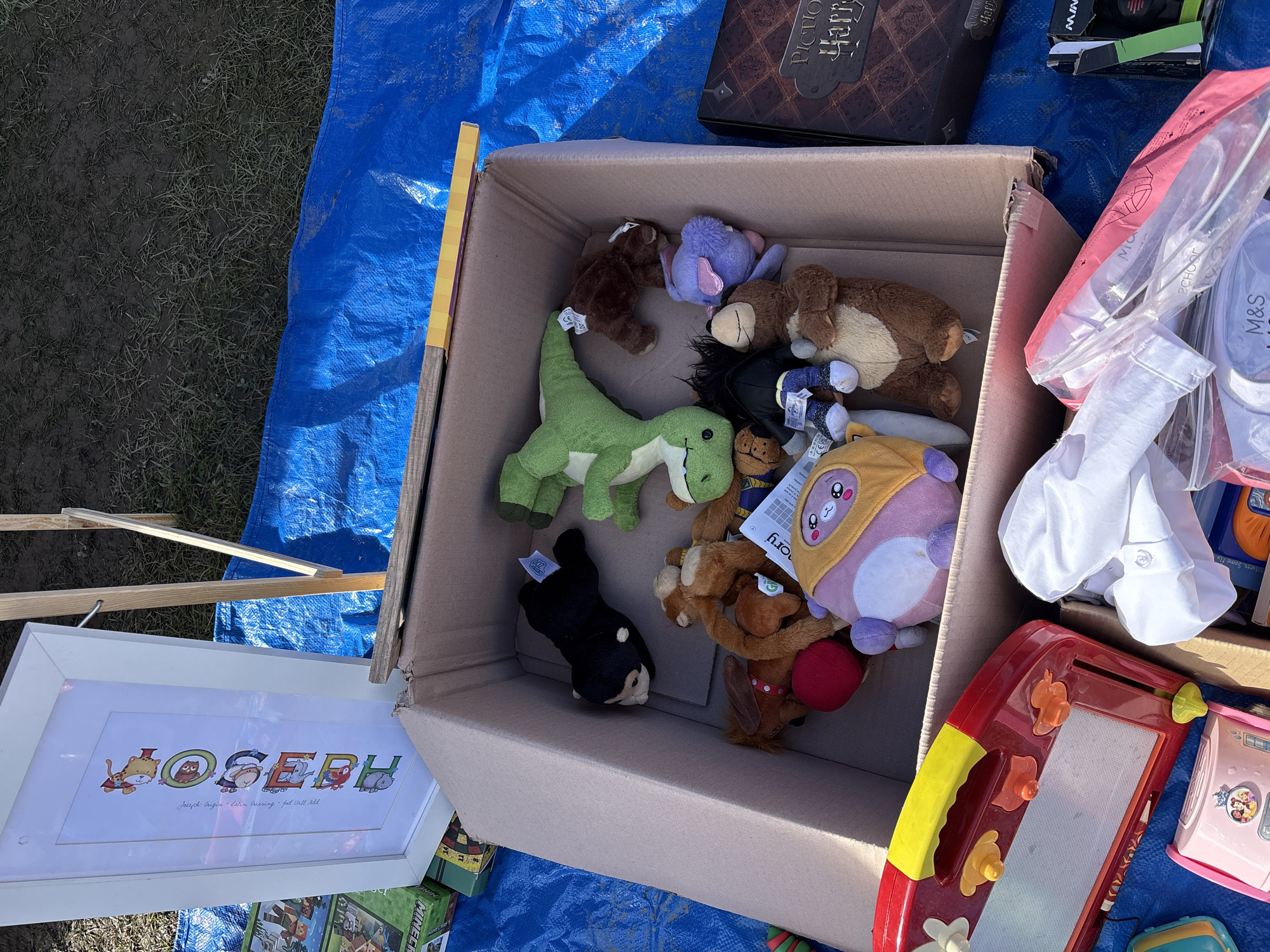 The image shows a collection of items arranged on a blue tarpaulin. Dominating the view is a cardboard box filled with stuffed animals, including teddy bears, a green dinosaur, and a black dog. Adjacent to the box is a framed picture with the name "Joseph" displayed in colorful, animal-themed letters. To the right of the box, there is a bag labeled "M&S SCHOOL," and several toys, including a red and yellow magnetic drawing board. The objects suggest a yard sale or flea market setting. Based on the OCR text in the image, it's highly likely that the image was taken in Chirk, United Kingdom.