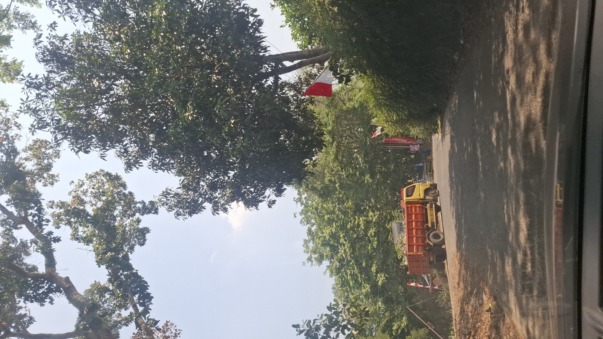 The image captures a scene on a paved road in Cibinong Hilir, Indonesia. A yellow dump truck with an orange bed, carrying a load of debris, is parked on the side of the road to the left of the frame. Behind the truck, several trees with lush green foliage line the roadside. A small Indonesian flag, red and white, is visible on a pole to the right of the truck, amidst dense green bushes. The sky is clear with a few wispy clouds. The foreground shows the dashboard and windshield of a vehicle, suggesting the photo was taken from inside a car. The road is partially shaded by the surrounding trees.