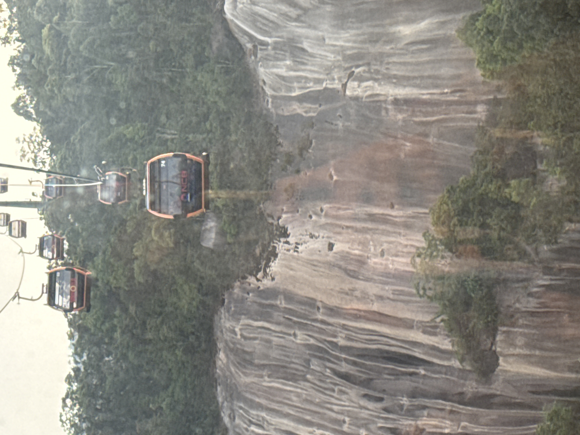 The image captures a scenic view of multiple cable car cabins traversing a mountainous landscape, characteristic of the Ba Na Hills in Đà Nẵng, Vietnam. The perspective is elevated, likely from within another cable car, looking down and across the route.
Several orange and black gondolas are visible, suspended by cables that stretch across the frame from left to right, implying movement. The most prominent cable car, positioned in the mid-right, clearly displays the letters "NCB" and the number "74" on its side. Other cabins follow in a line, receding into the background.
The left side of the frame is dominated by a dense, rich green tropical forest, covering the mountain slopes with lush vegetation. On the right, a massive, layered rock cliff face rises dramatically, showcasing intricate horizontal striations and rugged textures. Pockets of greenery cling to various parts of the cliff.
The atmosphere appears somewhat hazy or misty, contributing to a soft, diffused light across the scene. There are also visible circular marks, suggestive of raindrops or moisture on a window or camera lens, slightly blurring the image. The scene conveys the grandeur of the natural environment and the experience of aerial transport through it during daytime.