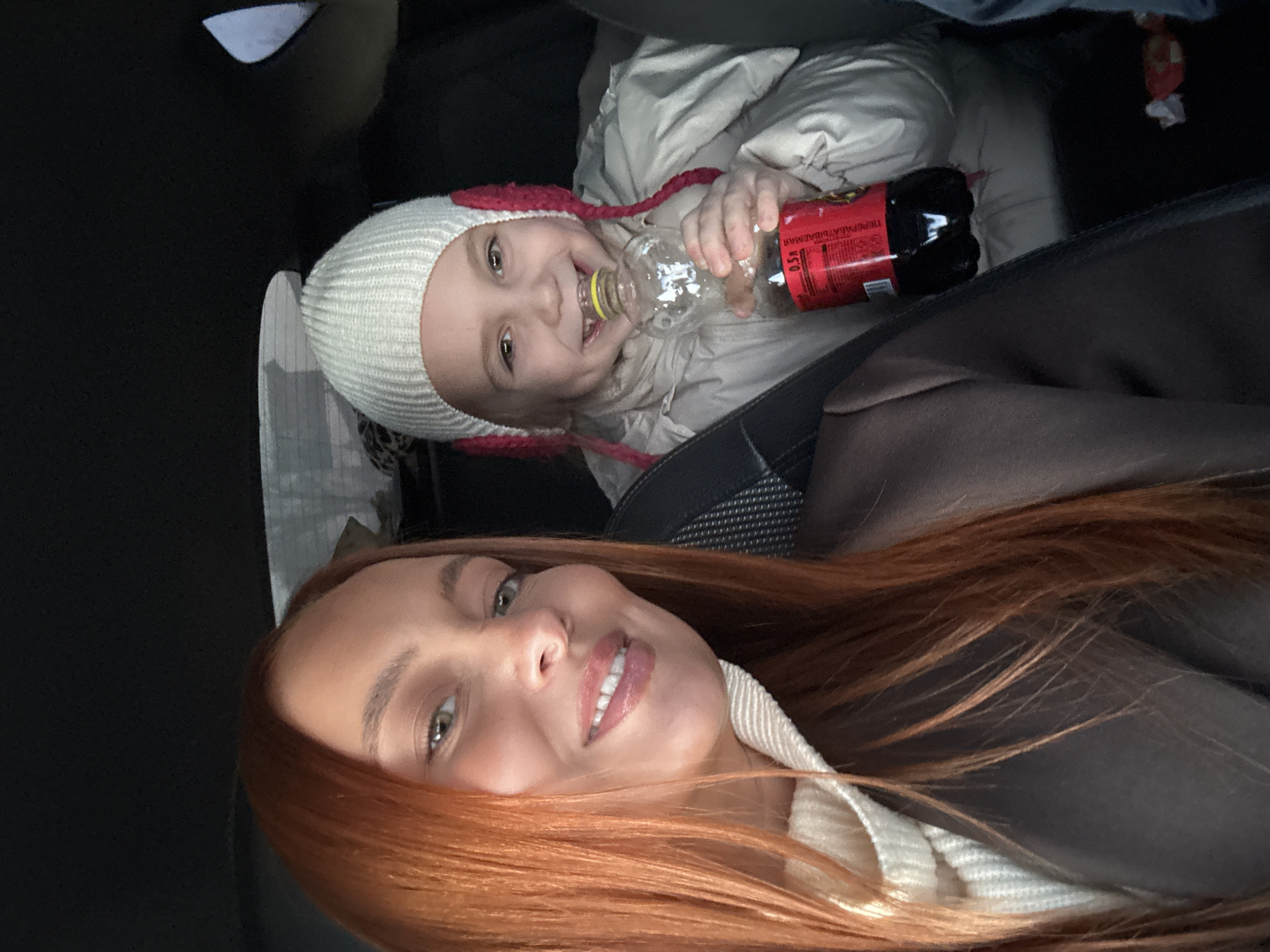 A selfie taken inside a car shows a woman with long, red hair smiling at the camera. Behind her is a young child wearing a white knitted hat with pink details and a light-colored puffer jacket. The child is holding a plastic bottle with a yellow cap and another bottle containing a dark liquid. The interior of the car is visible in the background, with a black car seat and other objects, including what appears to be a white cloth. The location appears to be Chaikovsky, Russia, due to the text on the darker bottle held by the child.