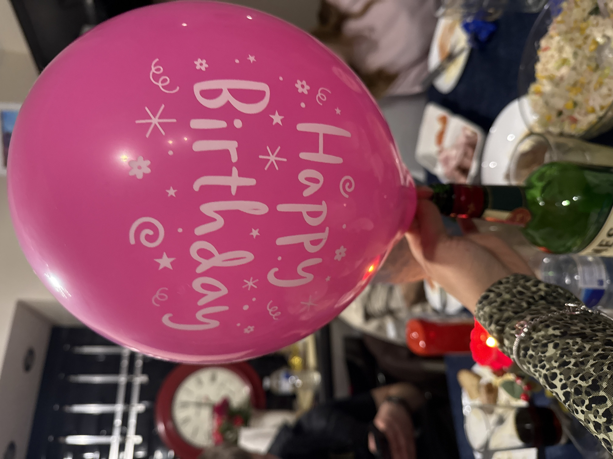 The image shows a pink "Happy Birthday" balloon being held in a room. The balloon is round with white lettering and celebratory symbols. In the background, there is a table laden with food, including a salad dish. People are seated around the table, though they are mostly out of focus. One person is wearing a leopard-print garment. There are wine bottles and glassware present, suggesting a celebratory meal. A red-framed clock is visible on a wall in the background. The location within Great Yarmouth, United Kingdom is not discernible from the image.