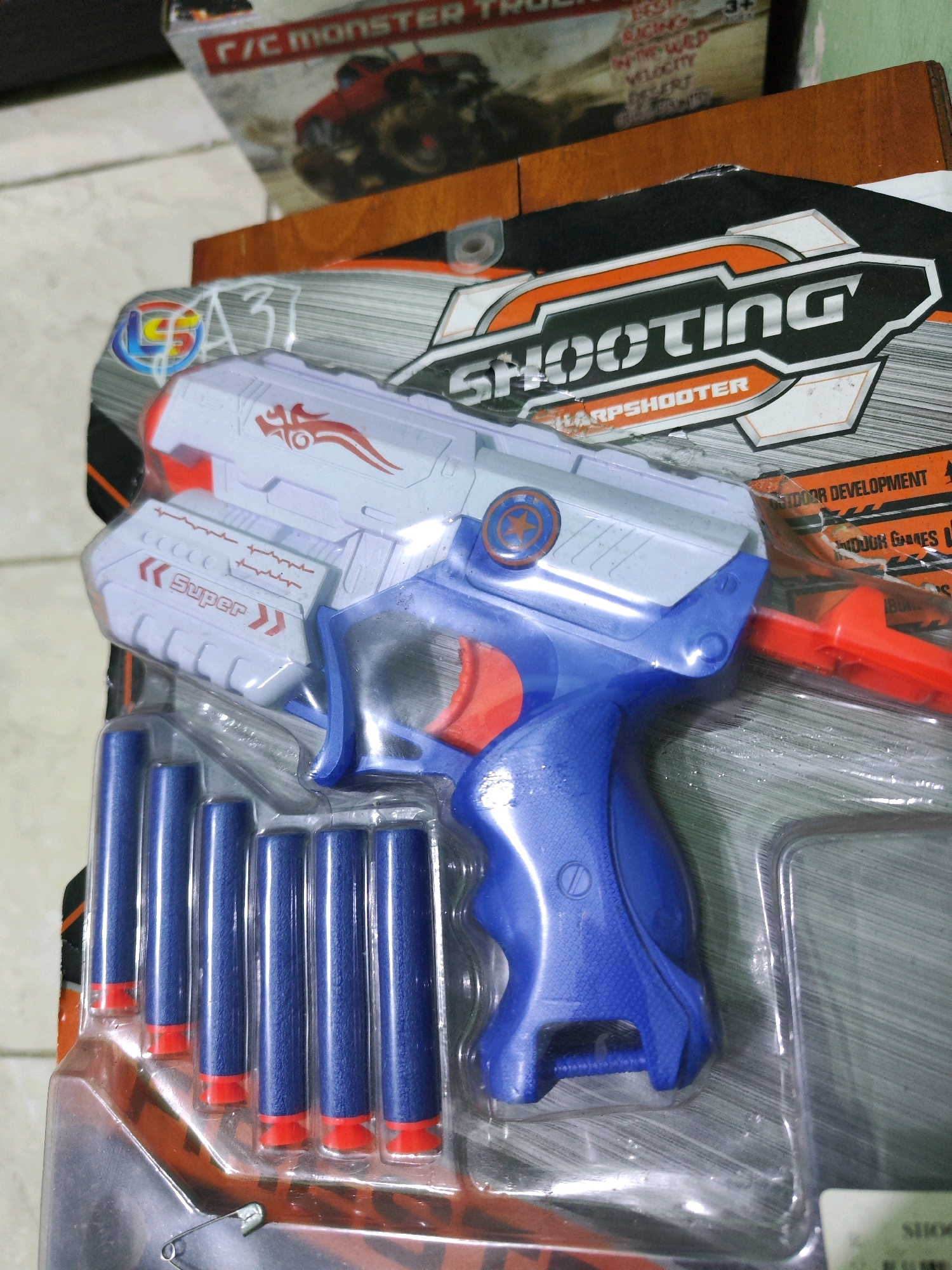 The image shows a close-up, angled view of a toy gun in its packaging, likely for sale in a store. The toy gun is primarily blue and white with red accents. It comes with several blue foam darts with red tips, arranged in a clear plastic blister pack. The packaging also features graphics, including the word "SHOOTING" in large, bold lettering and "HARPSHOOTER" below it. There are also other text elements and graphics on the packaging, some of which are partially obscured or out of focus. The background suggests a retail environment, with other products or packaging visible. The scene is likely indoors, given the lighting and the nature of the product. The specific location in Sroyo, Indonesia, cannot be determined from the visual information provided in the image.