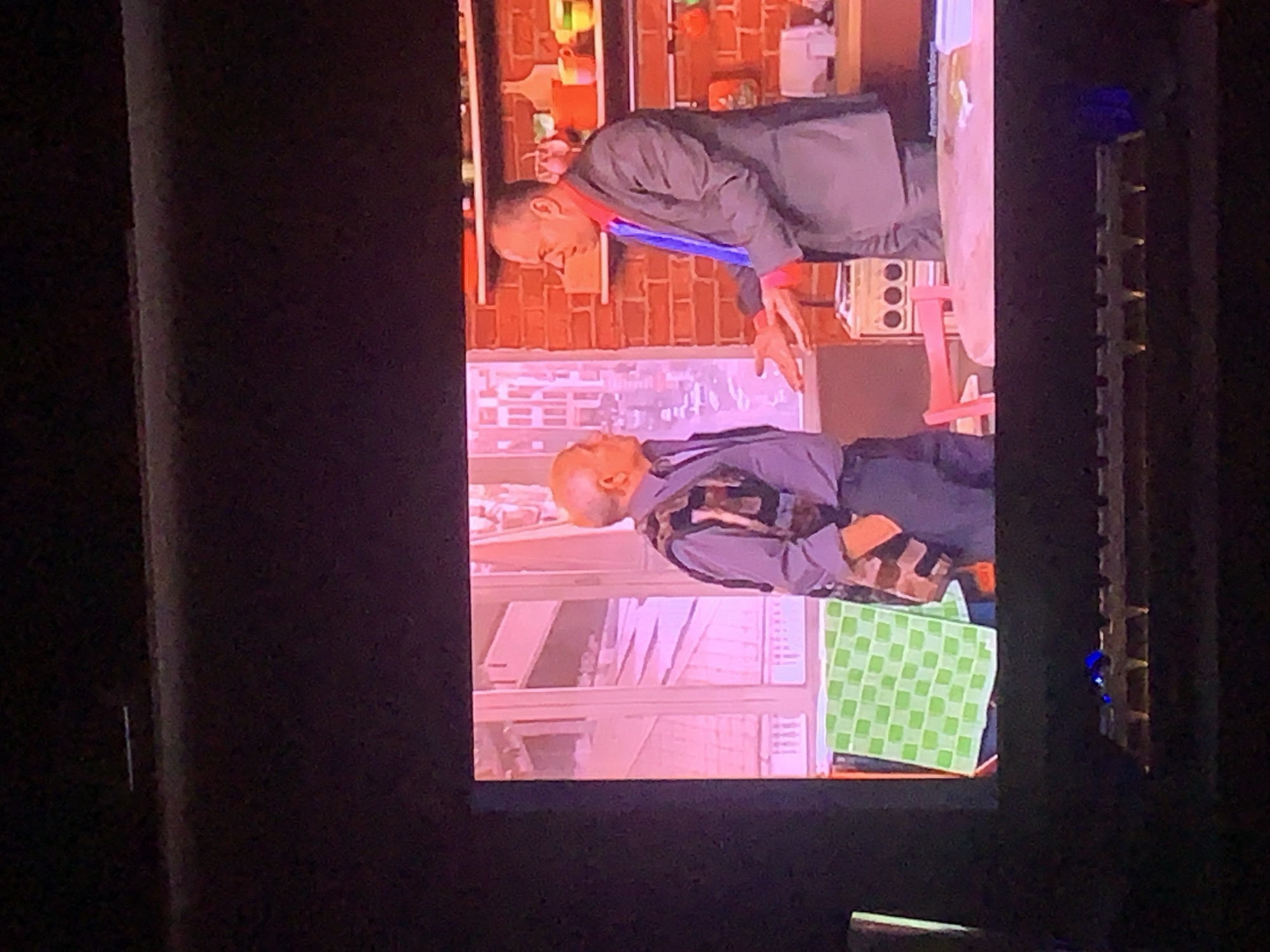 The image displays a scene from a television screen, depicting two men in what appears to be a studio set resembling a kitchen or living area. The man on the left is older, balding, and wearing a collared shirt and a patterned vest. He is looking towards the man on the right. The man on the right is younger, wearing a suit with a red shirt and blue tie. He is speaking with his hands outstretched, gesturing towards the man on the left. In the background, there is a brick wall, shelves with various items including mugs and kitchen utensils, and a window with a pinkish hue. A green checkered cloth is visible on the left side of the image, and a pink chair is in the foreground. The image is captured from a distance, with the television screen reflecting the surrounding environment. There is no indication of the city of Norilsk, Russia, or any specific geographical location.
