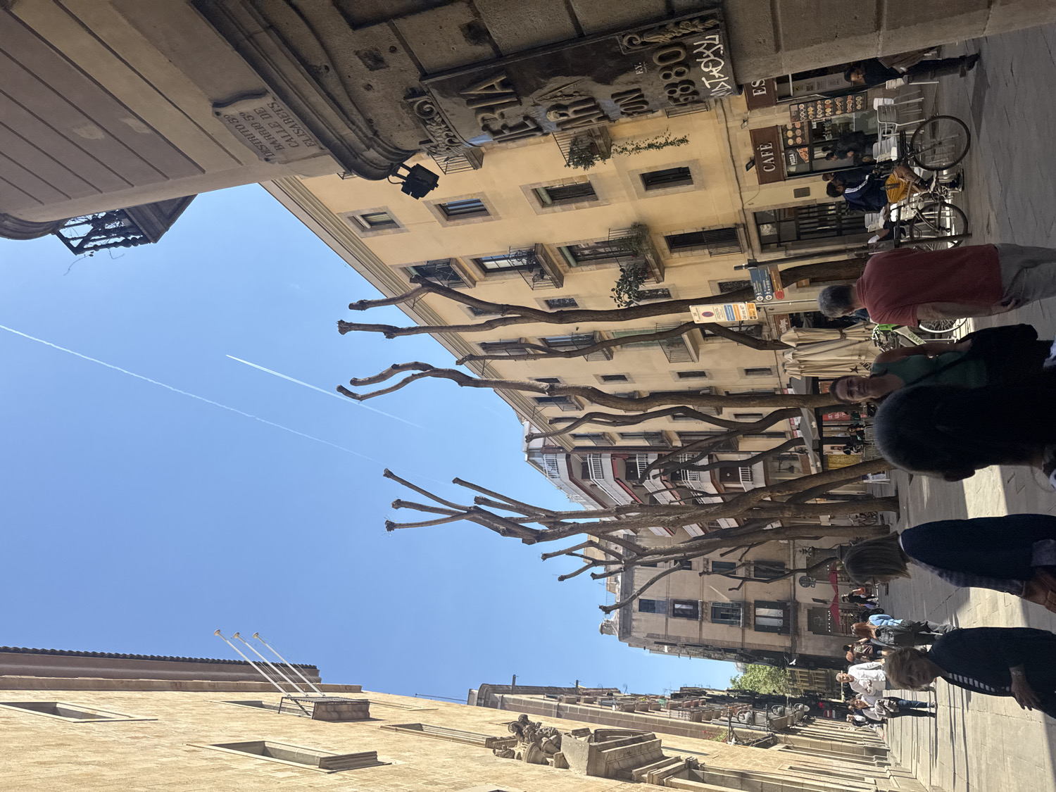 This is a bright, sunlit street scene in a bustling urban environment, likely Barcelona, Spain, given the provided context. The view looks upwards along a narrow street lined with multi-story, classic European buildings of light stone or plaster. The sky is a vivid, clear blue, traversed by two distinct white contrails from airplanes, suggesting good visibility and an active airspace.
Along the street, several bare-branched trees are visible, their gnarled forms reaching towards the sky, indicating late autumn, winter, or early spring. People are engaged in various activities: walking, standing, and potentially sitting at outdoor cafe tables, though these are mostly out of frame or obscured. Some individuals are clearly visible, dressed in casual attire, while others appear as blurred figures, suggesting movement. Bicycles are parked near a cafe.
Notable architectural details include ornate balconies on some buildings and commercial signs. Visible text includes a prominent street sign reading "DISTRITO 39," "BARRIO S," and "CALLE DE ELISABETS," confirming the Spanish setting. Other readable signs include "CAFÈ," "PIZZA," "FONTS JEYA" (partially), and "ES." Numbers like "1880" are also discernible on some signage. The overall impression is one of a lively pedestrian street during a clear, sunny day.