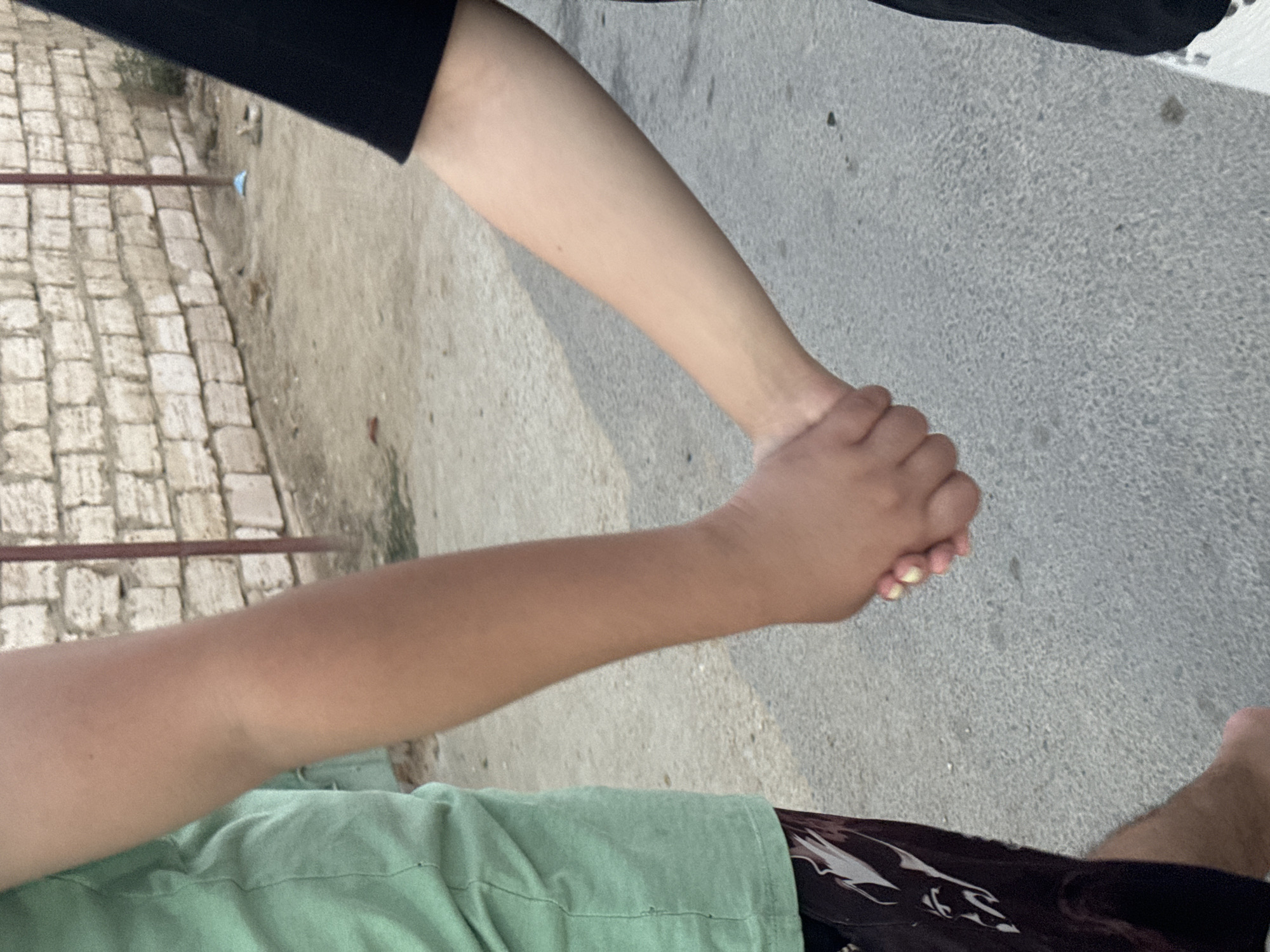 The image shows a close-up of two people's arms being held, with a brick wall in the background and a paved ground surface. The person on the left is wearing a green t-shirt and patterned shorts. The person on the right is wearing a black t-shirt. Their hands are clasped together. The environment appears to be outdoors, possibly a street or walkway in a city, as indicated by the paved surface and the brick wall construction. The lighting suggests it is daytime. The location is Atameken, Kazakhstan.
