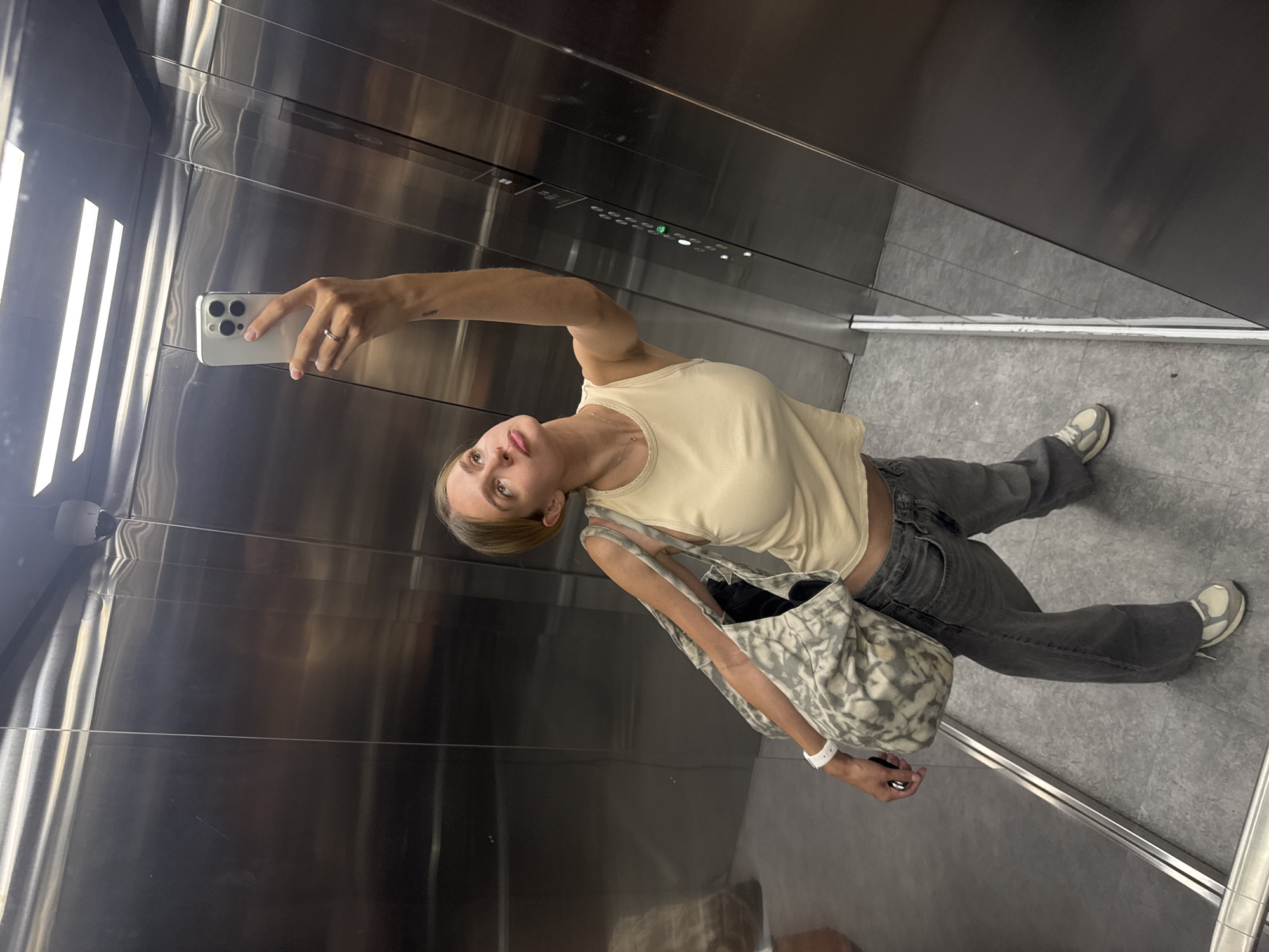 A young woman is taking a selfie inside a stainless steel elevator. She is wearing a beige tank top, grey jeans, and grey and white sneakers. She has a large patterned tote bag slung over her shoulder and is holding a white smartphone in her right hand to take the picture. Her left hand is partially visible, adorned with a ring and a white wristband. The elevator interior consists of brushed steel walls and a tiled floor. The elevator panel with buttons is visible above the woman. The exact city and location are unknown.
