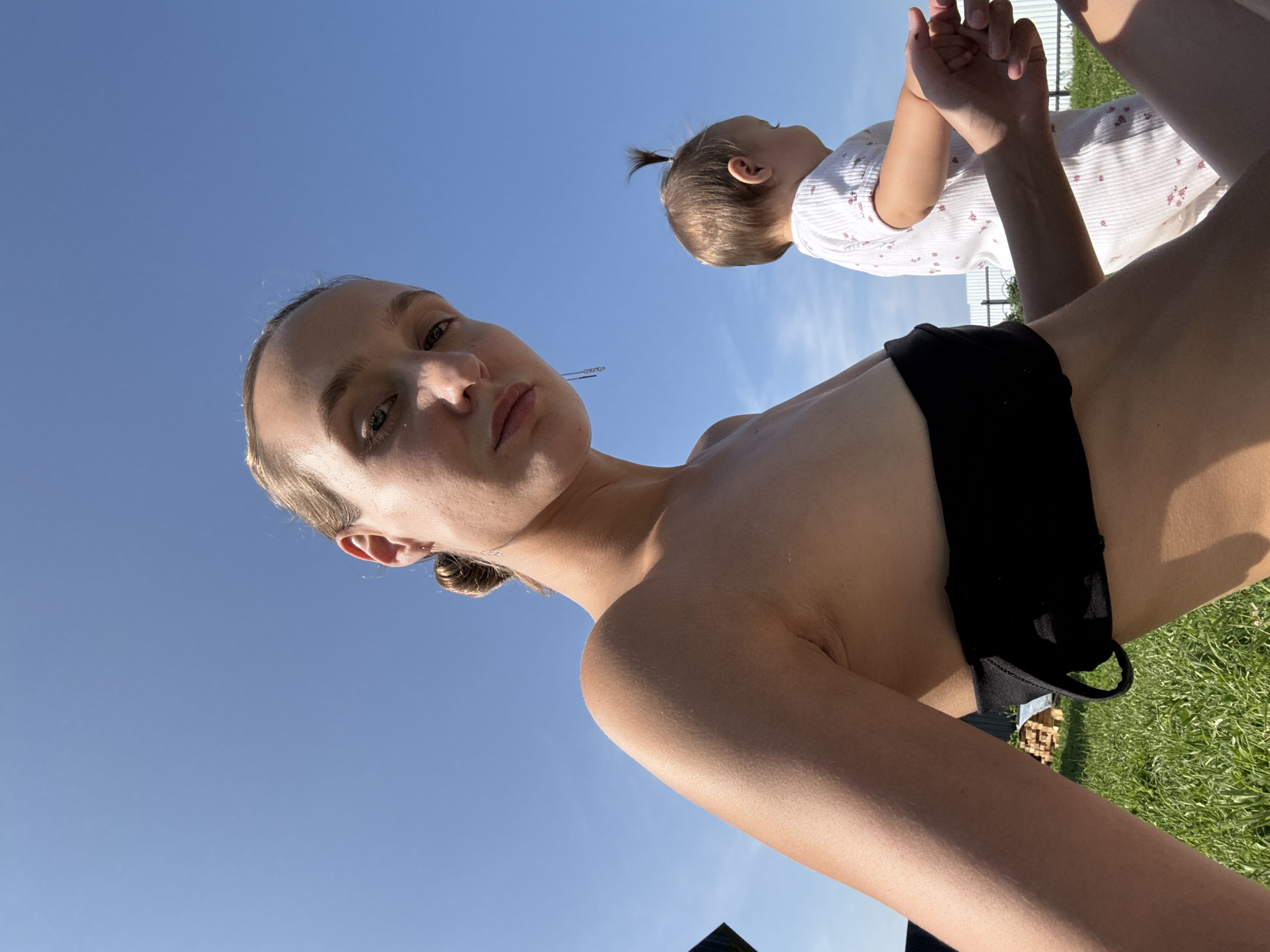 A low-angle, medium shot captures a woman and a child outdoors under a clear blue sky. The woman, with her blonde hair pulled back, is in the foreground, looking directly at the camera. She is wearing a black strapless top. To her right and slightly behind her, a young child in a white onesie with a floral pattern is visible from the back. The child has a small ponytail. They are positioned on a grassy area, with what appears to be a small wooden structure and a fence in the background. The setting suggests a rural or suburban environment, possibly in Podshivalovo, Russia. The lighting indicates it is daytime, likely sunny.