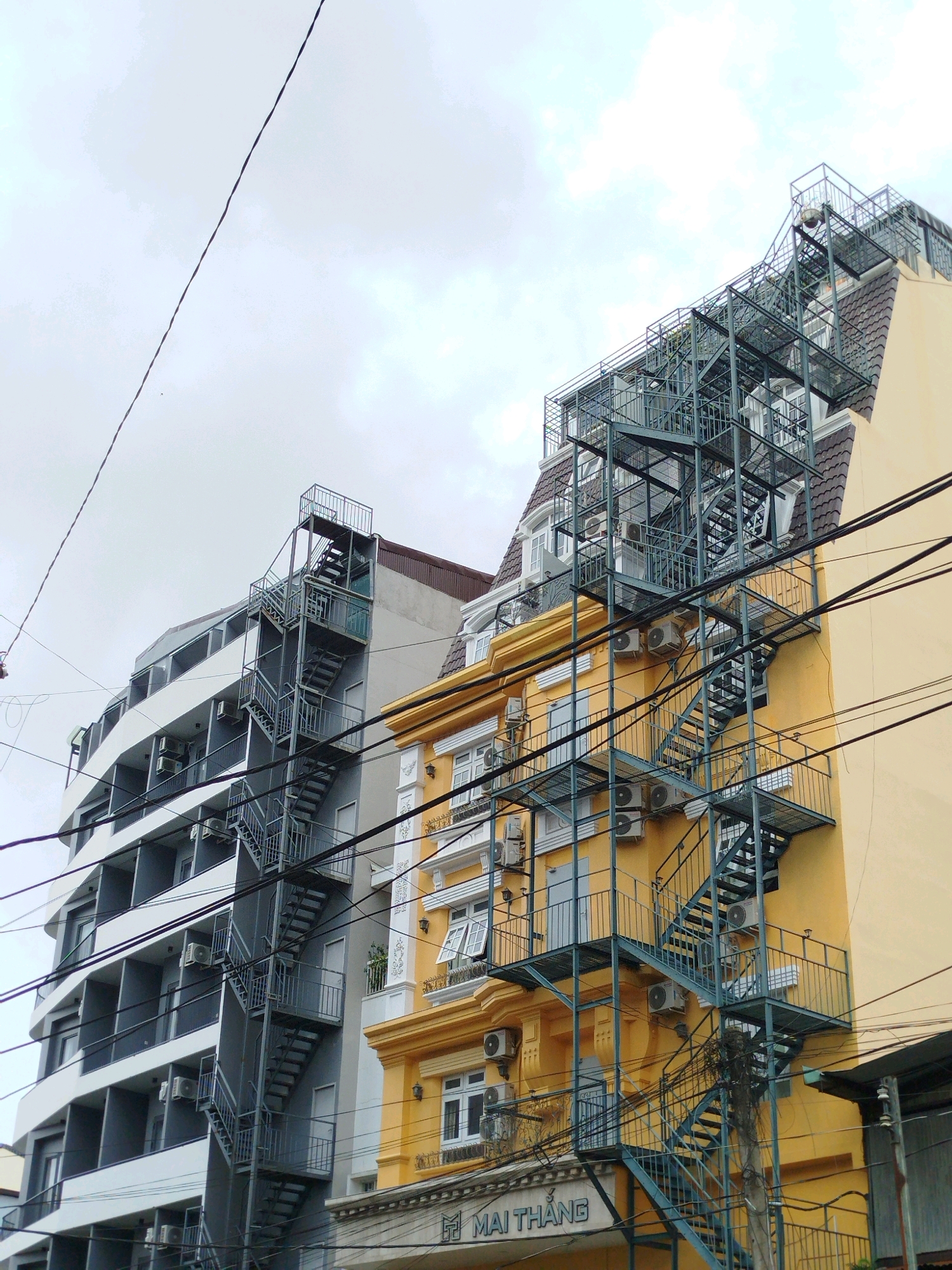 The image depicts an overcast day in Da Lat, Vietnam, showcasing a street scene dominated by buildings and utility wires. Two distinct buildings are prominently featured. On the left, a modern, multi-story building with a white and gray facade features numerous balconies and visible air conditioning units. An external metal staircase runs vertically along its side. To the right, a narrower, older building with a yellow facade displays architectural details like decorative molding around windows and a mansard-style roof. This building also has an extensive external metal staircase that appears to be a fire escape, serving both buildings and extending over utility wires. The sky is a uniform gray, suggesting cloudy weather. Numerous black electrical wires crisscross the upper portion of the frame, obscuring parts of the buildings and sky. The overall impression is one of urban density and infrastructure. No people are visible in the frame.