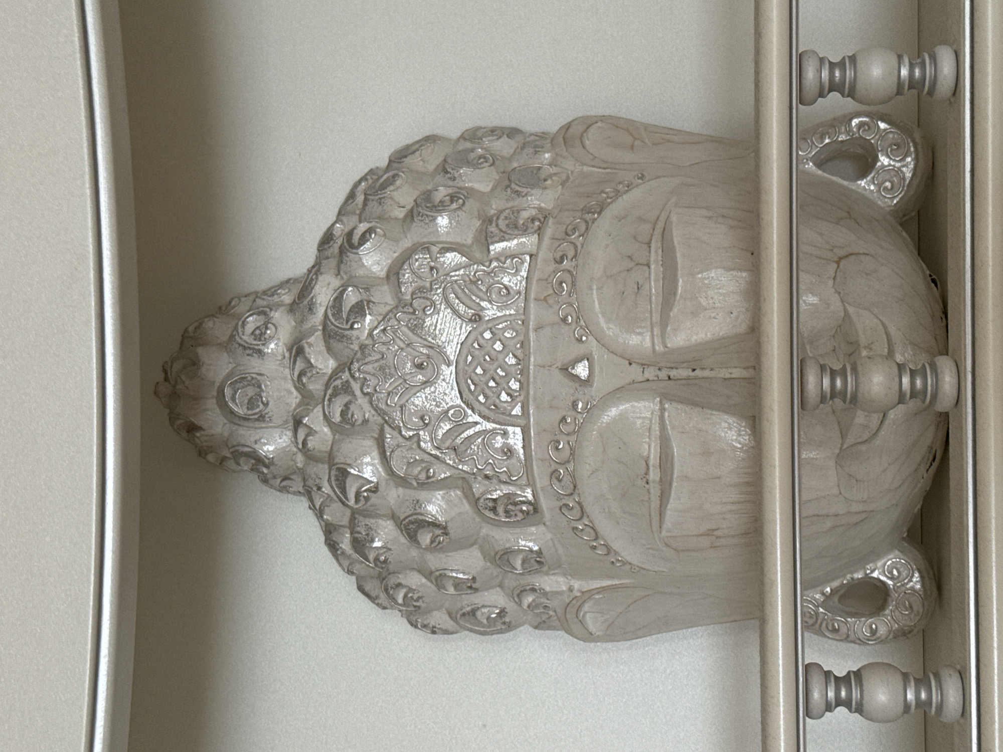 A decorative, pale, sculpted head of Buddha rests in a narrow shelf or display space. The head is intricately detailed, with rows of small rounded curls covering the top and back of the head. There's an ornate headband above the forehead, and the eyes are closed in peaceful repose. The face has a textured, aged appearance. The display space is painted white, and a portion of a vertical post or frame is visible on the right side, obstructing part of the head. There is no evidence that this image was taken in Vsevolozhsk, Russia.