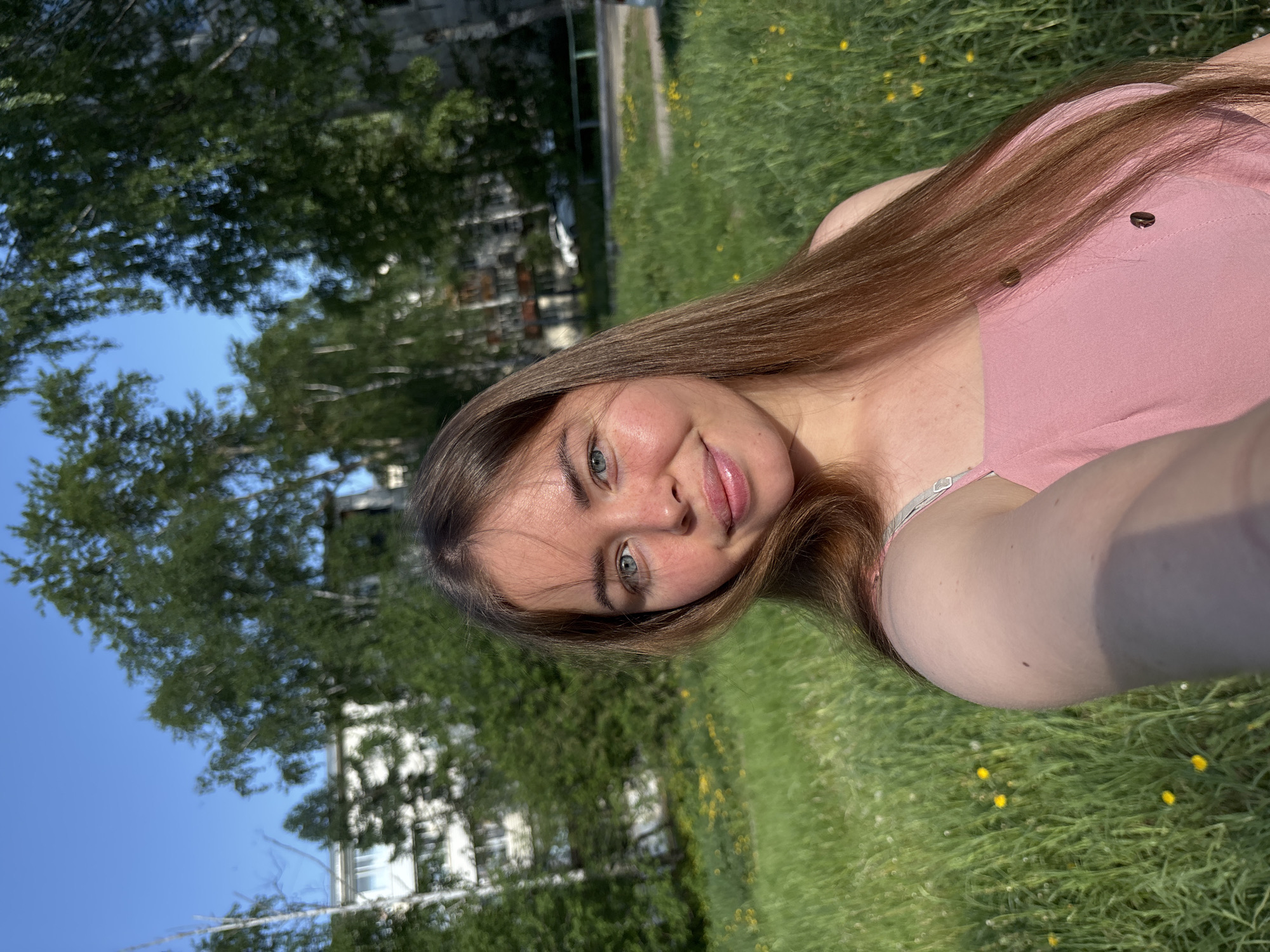 A young woman with long, straight brown hair is taking a selfie outdoors. She is wearing a pink top and has light skin and blue eyes. The background includes a grassy field dotted with small yellow flowers, trees with green foliage, a building, and a clear blue sky. Based on the information available, this scene is likely located in a park or green space in Izhevsk, Russia.