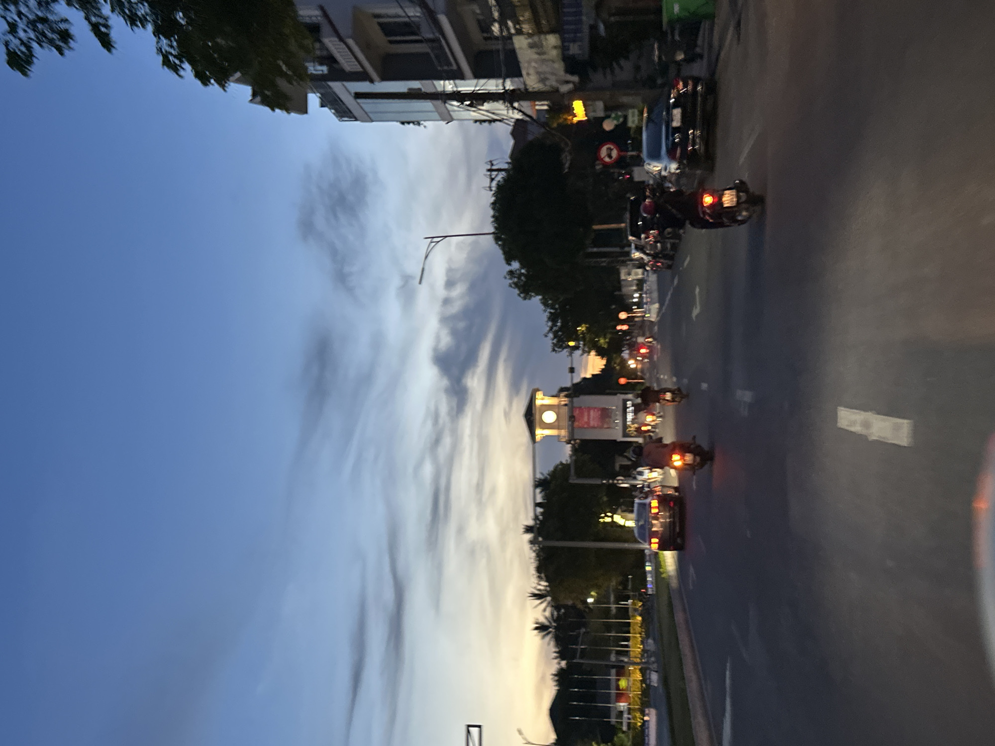 The image depicts a street scene at dusk in Da Nang, Vietnam, likely near the Dragon Bridge or Han River. The sky is a gradient of deep blue to orange, indicating sunset or twilight. A prominent clock tower with illuminated faces stands in the middle ground, suggesting it's a landmark. The street is moderately busy with vehicular traffic, including cars and numerous motorbikes, some carrying passengers. One motorbike in the foreground has its brake lights illuminated. Buildings line the right side of the street, with a multi-story structure featuring balconies and glass facades. Trees are visible along the left side of the road, with palm trees suggesting a tropical or subtropical location. The road itself has visible lane markings.