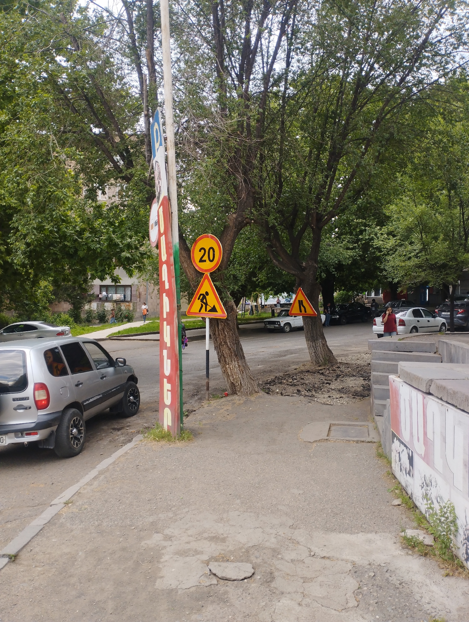 The image depicts a bright, daytime street scene, likely in an urban or suburban setting, consistent with Yeghegnadzor, Armenia. In the foreground, a cracked and worn paved sidewalk is visible. On the left, a silver SUV, resembling a Chevrolet Niva, is parked partially on the sidewalk. To the right, a low concrete wall with faded painted text, including "ՕԼԻ" and other illegible parts, borders the path.
A cluster of large trees with dense green foliage dominates the midground, with some directly on the street edge. Attached to one tree trunk are two prominent traffic signs: a yellow circular sign displaying "20," indicating a speed limit, and below it, a yellow triangular sign depicting a person shoveling, signaling road work ahead. Further down the street, another tree has a yellow triangular sign showing converging lines, signifying a road narrows.
Several cars are parked along the street beneath the trees, including white and dark-colored sedans. A woman in a red top is visible walking on the right side of the street near the parked cars, while other pedestrians are in the distance on the left sidewalk. Buildings are partially visible through the trees in the background. A tall, white pole with a red-painted side stands near the silver SUV, featuring partially visible Armenian text, "ՆԱՐԱՆԳ," likely part of a local business sign. The scene indicates ongoing road work in an active yet not overly congested street environment.