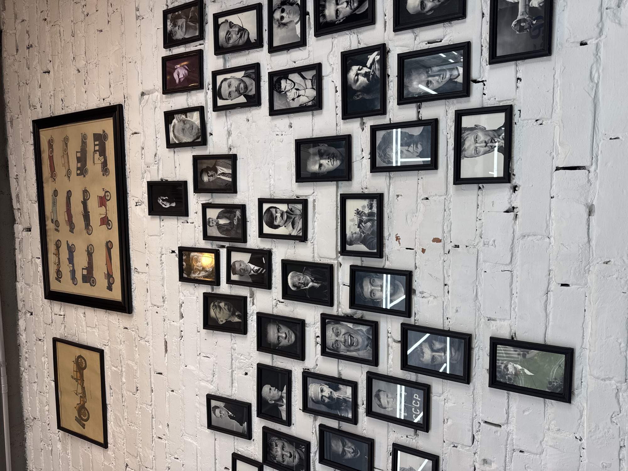 A white brick wall displays a collection of framed pictures and posters. On the left, two framed posters depicting antique cars hang vertically. To the right, a series of smaller framed pictures, predominantly black and white portraits, are arranged across the wall. The scene appears to be indoors. While specific details to confirm it is in Novosibirsk, Russia are lacking, the "CCCP" text on one of the picture frame could indicate a Soviet or post-Soviet context, but it is not a definitive identifier of the location.