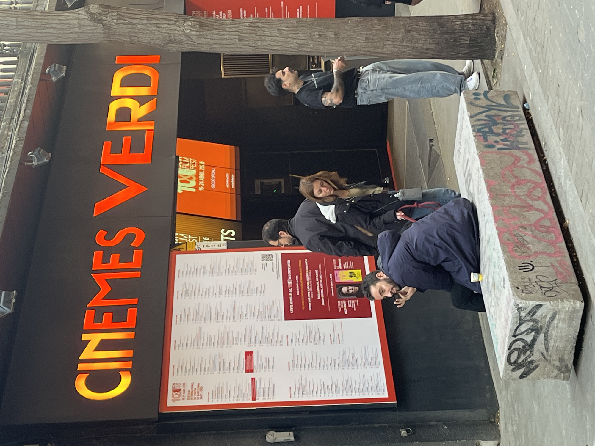 The image captures the facade of the "Cinemes Verdi" in Barcelona, Spain. A large orange sign spells out the cinema's name. Below the sign is a poster advertising the "100 Film Fest" from April 15-24, 2016. A second, larger poster displays a list of film titles. Several people are in the scene. One man stands looking up, a woman stands beside him and two other men stand on the sidewalk looking at the large poster. One is holding an object in his hand. A concrete bench with graffiti on its surface is in the foreground. A tree trunk stands near the entrance.