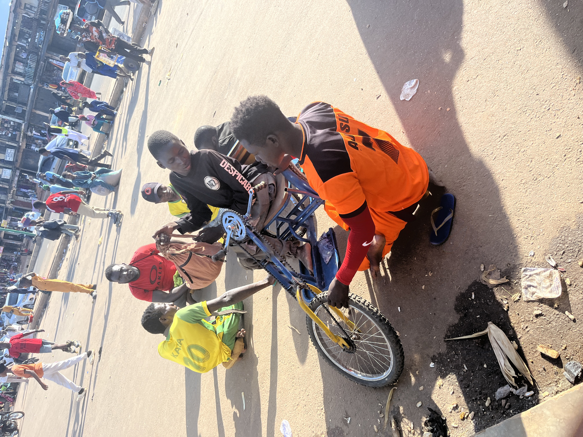 The image depicts a street scene in the city of Jos, Nigeria. In the foreground, a young Black male wearing a bright orange shirt with black accents is on his knees, seemingly working on a blue bicycle. He is positioned next to the bicycle's rear wheel, with his right hand near the chain and gears. To his left, another young Black male in a yellow shirt with the number '10' and green sleeves kneels on the ground, looking down. Behind them, a Black male wearing a red t-shirt sits with a patterned fabric bag on his lap. The bicycle has a small front wheel and a larger rear wheel, with blue metal framing. In the background, a bustling street is visible with various people walking and standing, some wearing traditional clothing. Vehicles, including cars and motorcycles, are present, and buildings line the street. The ground is paved with asphalt and shows signs of dirt and scattered debris, including what appears to be peanut shells. The lighting suggests it is daytime, with strong shadows cast on the ground.
