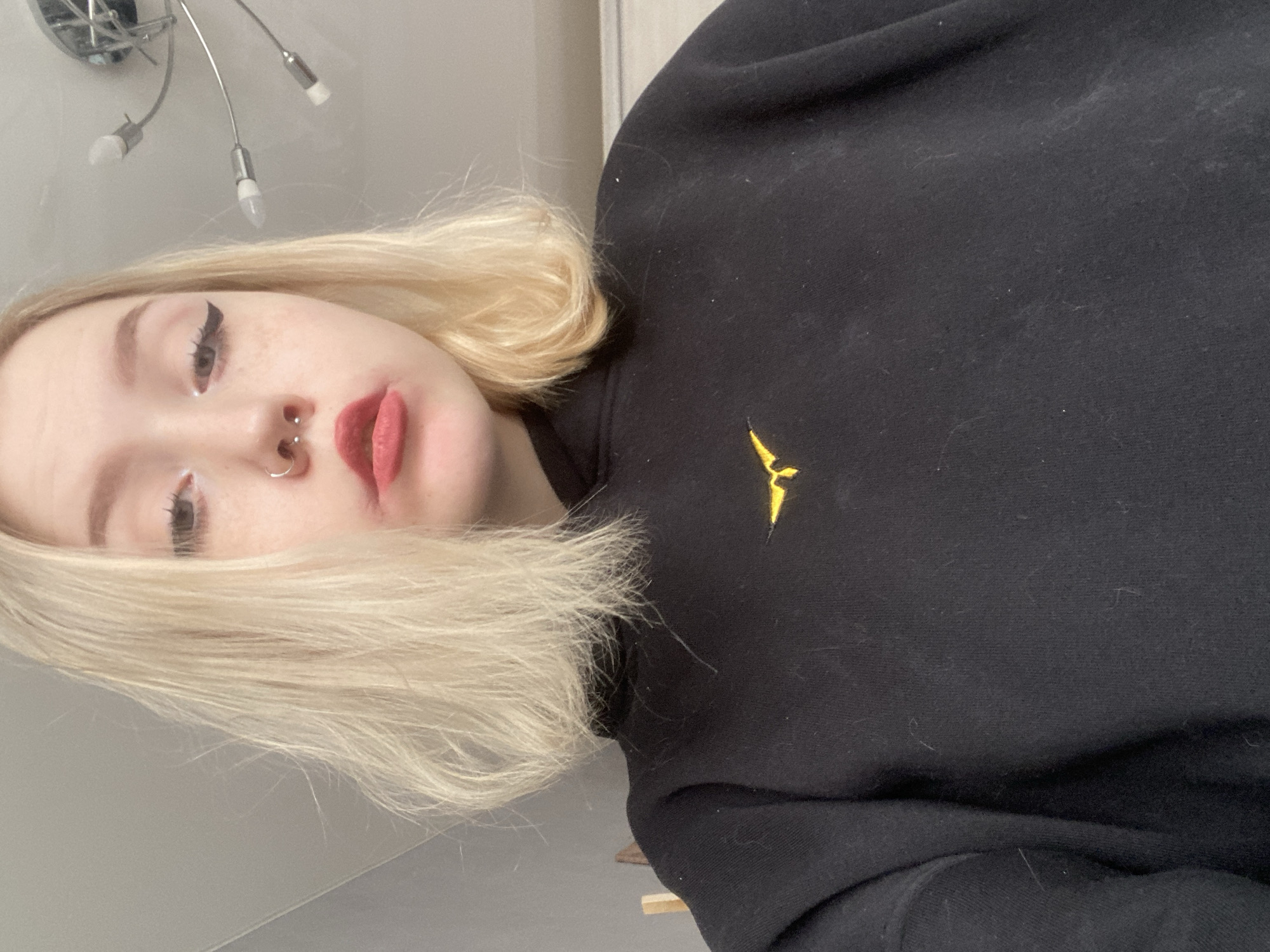 A selfie of a fair-skinned person with blonde hair and facial piercings (septum and nostril) is taken indoors. The person is wearing a black sweatshirt with a small, stylized yellow bird emblem on the chest. The background includes a light-colored wall and a portion of a modern-style ceiling light fixture. The person's eye makeup is visible, including eyeliner. The person has fair skin and freckles. The lips are red. The photo appears to be rotated counter-clockwise. The location is not inferable as Izhevsk, Russia.