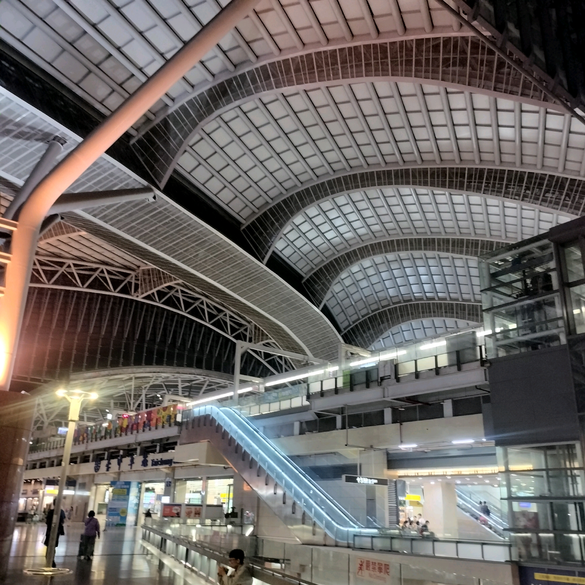 This image depicts the interior of Taichung Railway Station in Taichung, Taiwan. The station features a grand, arched ceiling constructed with white panels and a metal framework, illuminated by overhead lighting and the ambient glow from within the building. A prominent, illuminated escalator with blue light accents is visible, leading upwards to the station's upper levels. Various platforms, walkways, and glass-enclosed structures are present, indicating a busy transportation hub. Several individuals are captured in motion within the station, some walking with luggage, others ascending or descending the escalator, and a few gathered on platforms. Signs and decorative elements are visible along the upper levels, suggesting areas for shops or waiting rooms. The overall scene conveys a modern and spacious architectural design characteristic of a contemporary railway station.