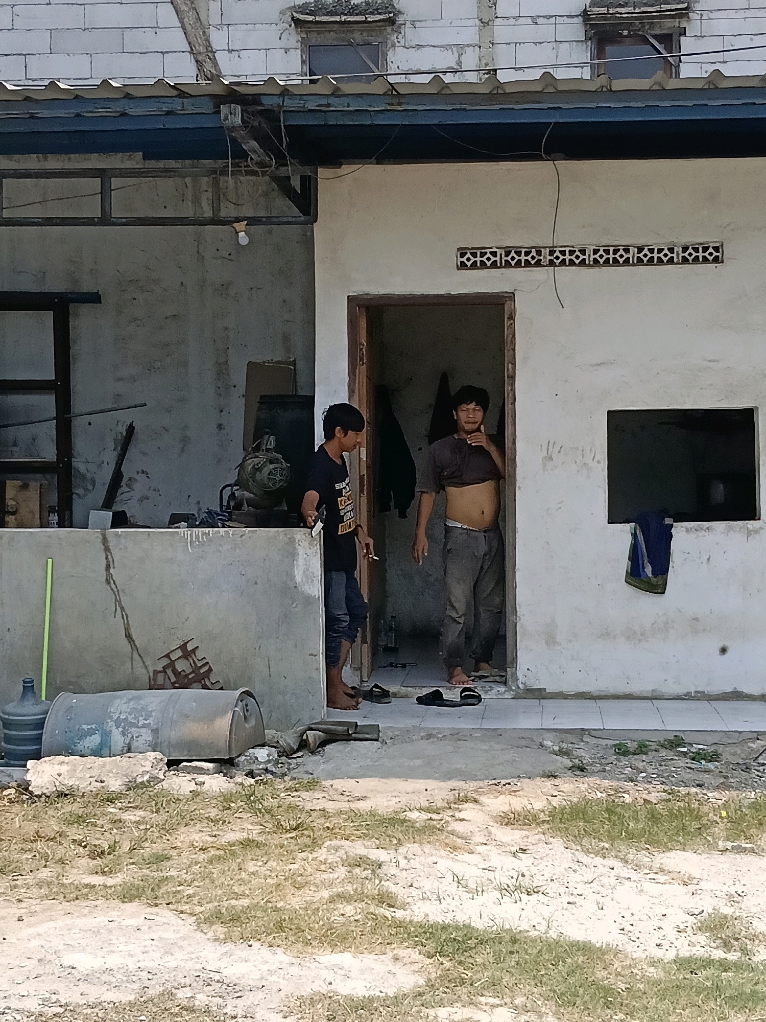 In the city of Belimbing, Indonesia, a medium-shot captures two young men standing near an outdoor structure. The man on the left, with short black hair, is wearing a black t-shirt and dark pants, and is seen holding a cigarette. He is standing next to a concrete counter which displays various tools and equipment, including what appears to be a motor and a metal pipe. The man on the right, also with short black hair, is wearing a cropped brown t-shirt and grey pants, revealing his midriff. He stands in an open doorway. The building features a corrugated metal roof and white walls with a decorative vent. In the foreground, a large plastic water jug and a metal barrel are visible on the dusty ground, with dry grass scattered around. The scene appears to be an outdoor workshop or storage area.